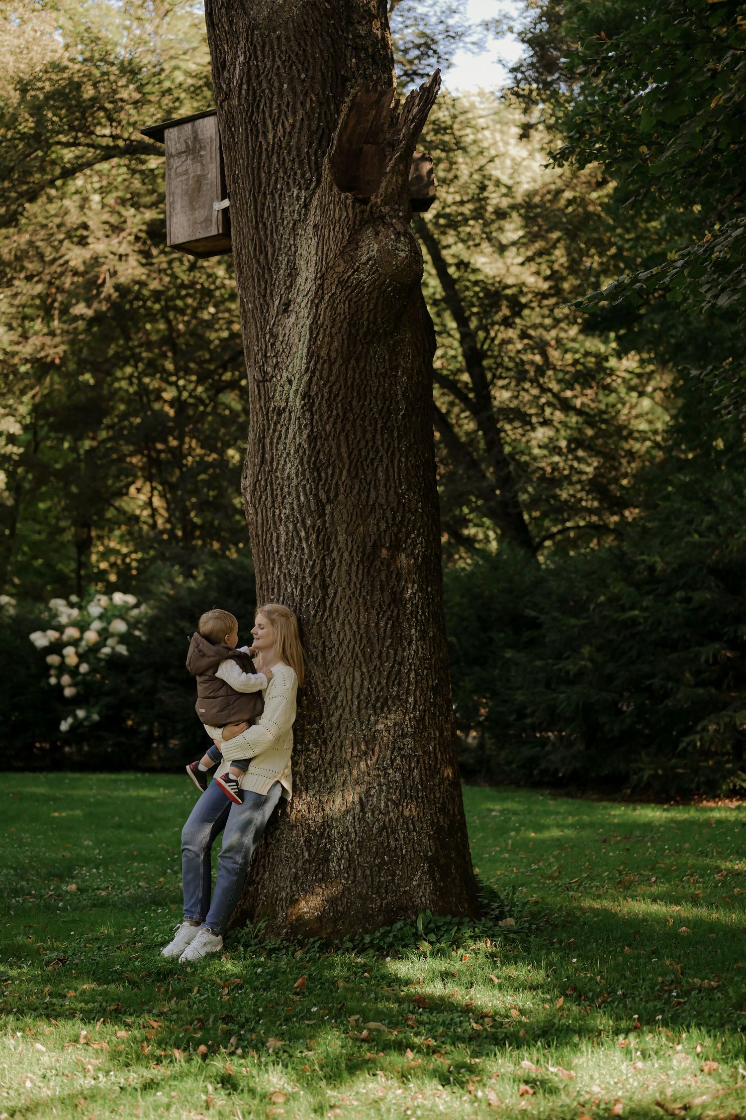 A mother holds her young child while leaning against a large tree trunk in a lush green park.