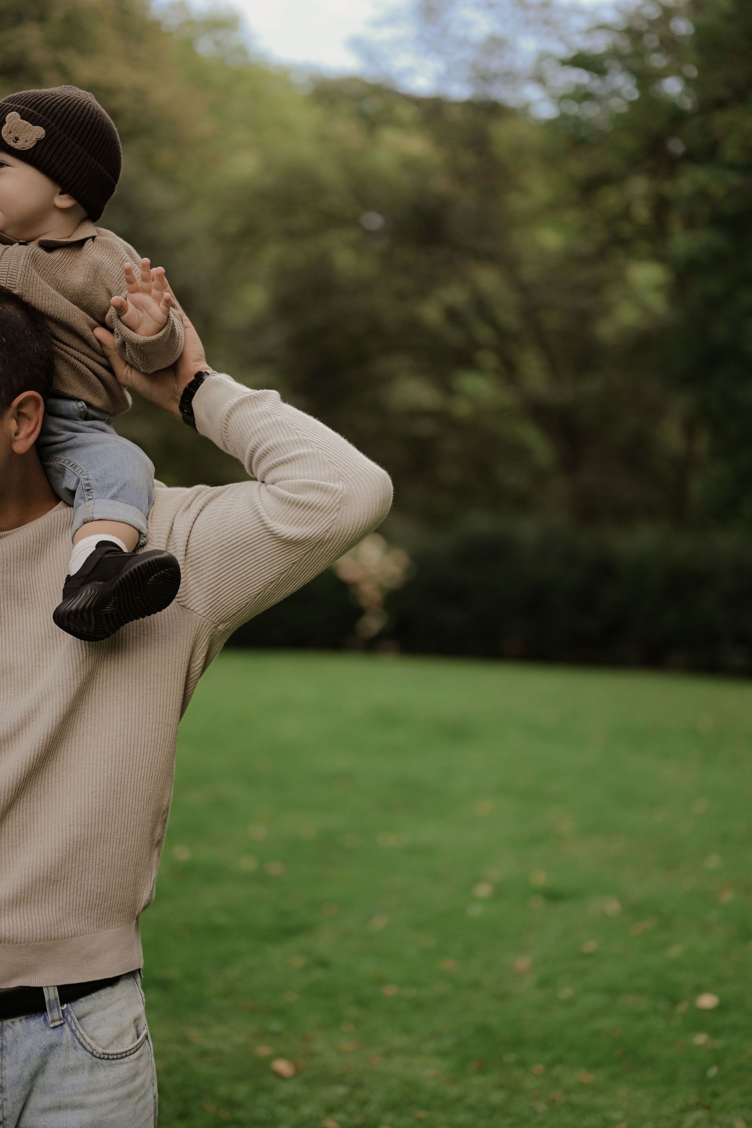 A parent lifting a baby on their shoulders in a lush green park, both wearing casual warm clothing.