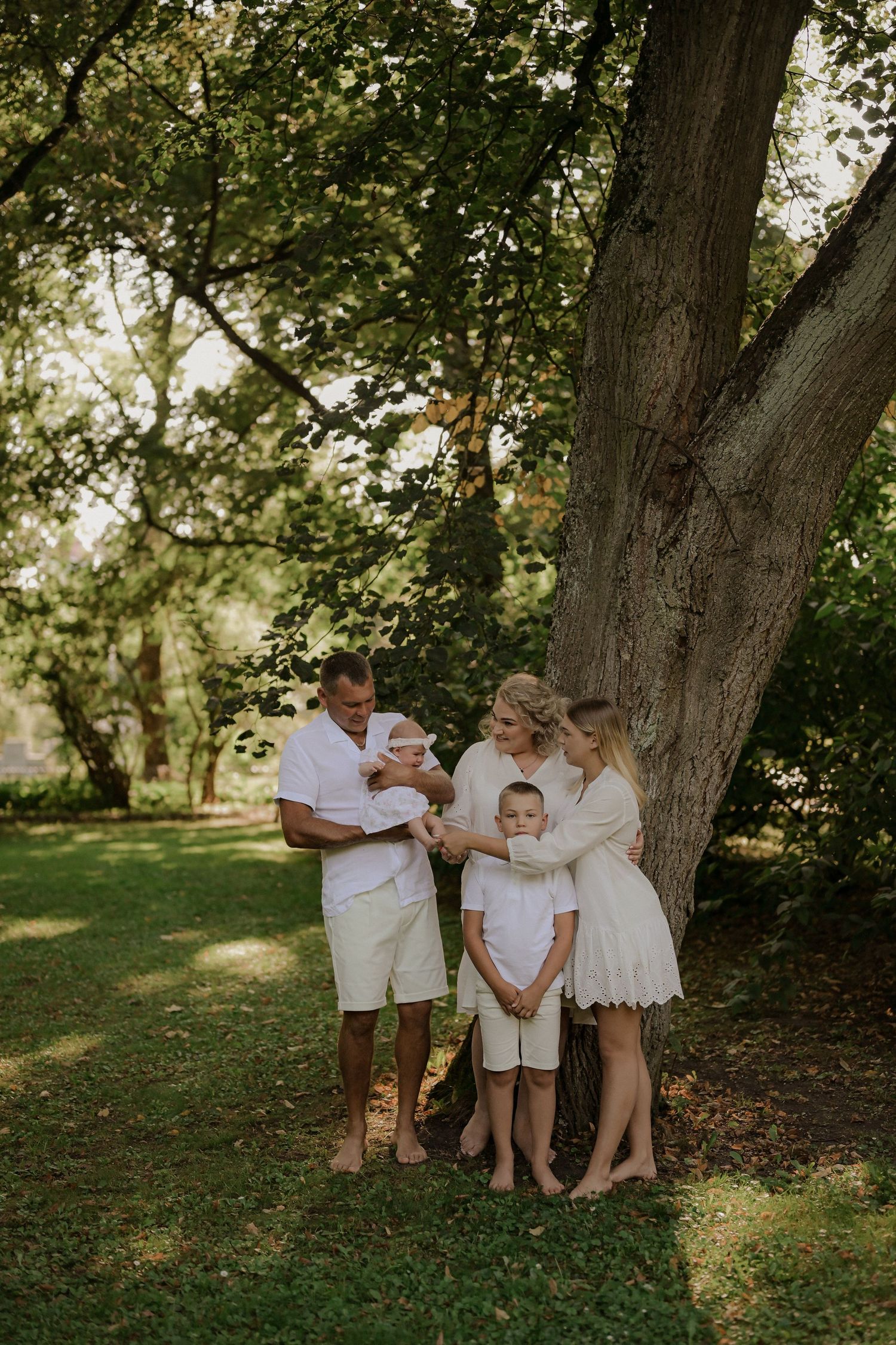 Family of four in white outfits standing barefoot under a large tree in a lush green park setting.