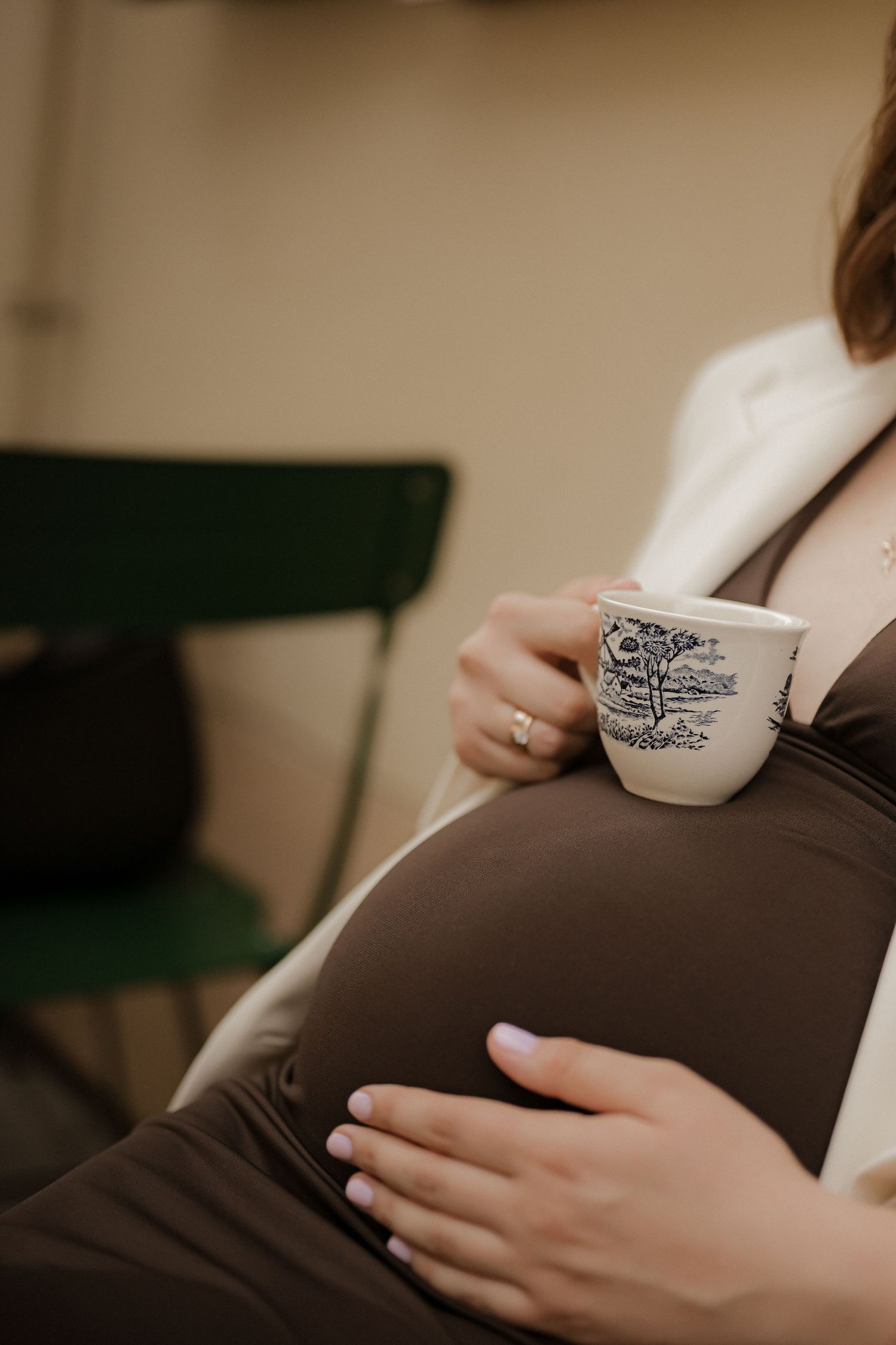 Pregnant woman in brown dress holding decorative teacup while cradling baby bump, seated in cozy cafe setting.