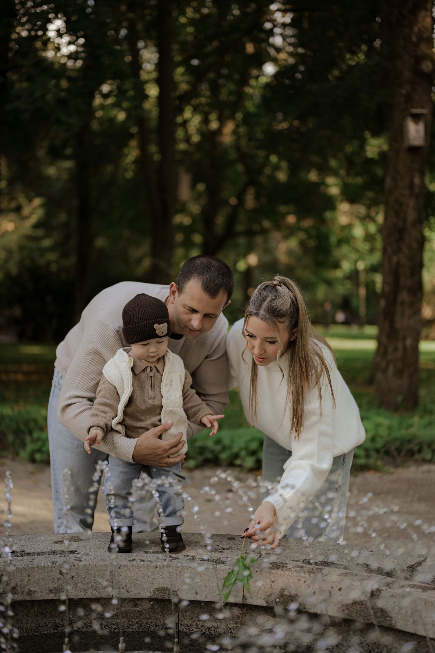 A young family with a toddler enjoys a park fountain, parents leaning in as child reaches toward splashing water.