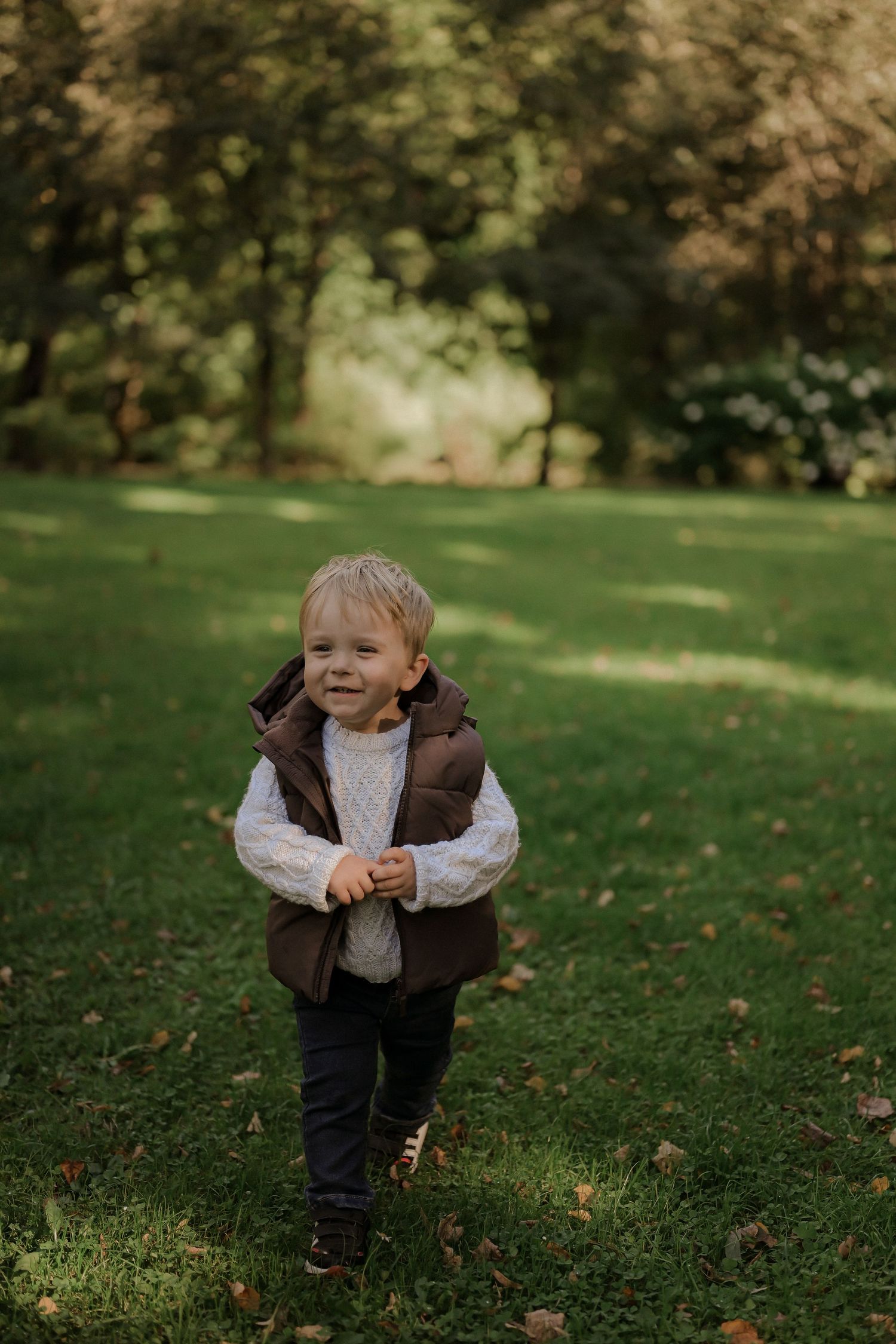 Toddler boy in brown vest and white sweater walking on green grass in a park during autumn.