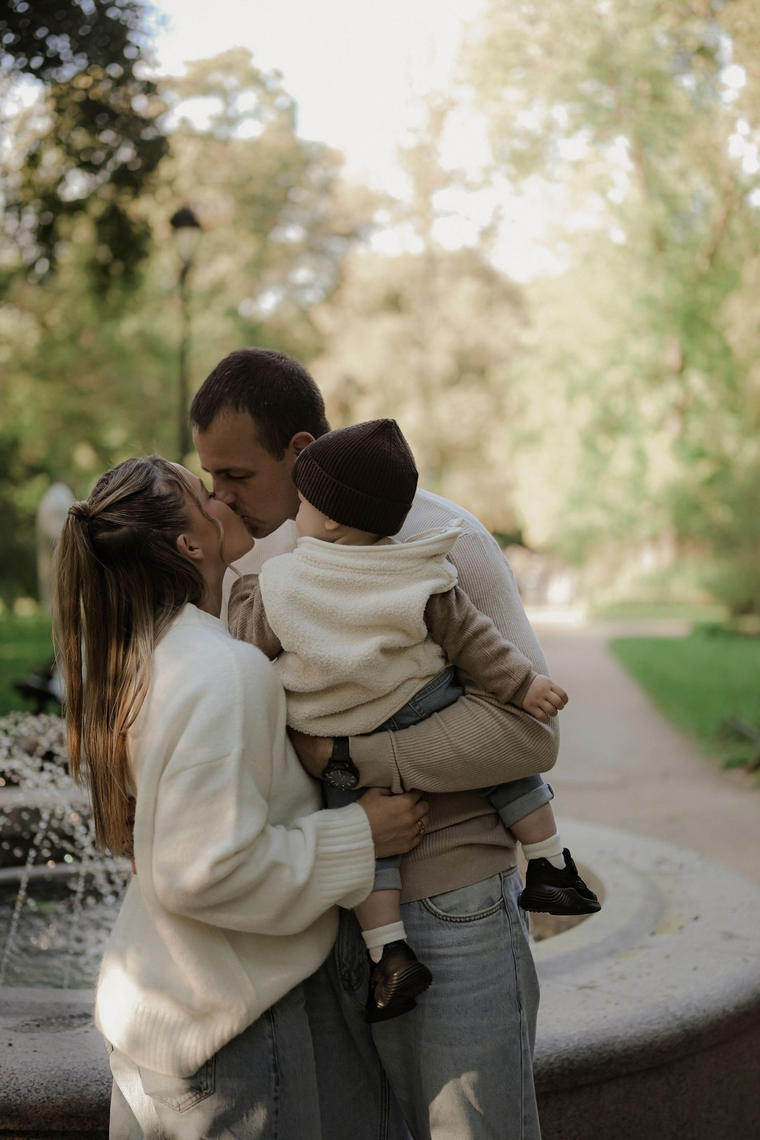 A couple sharing a kiss while holding their baby in a lush green park near a fountain.