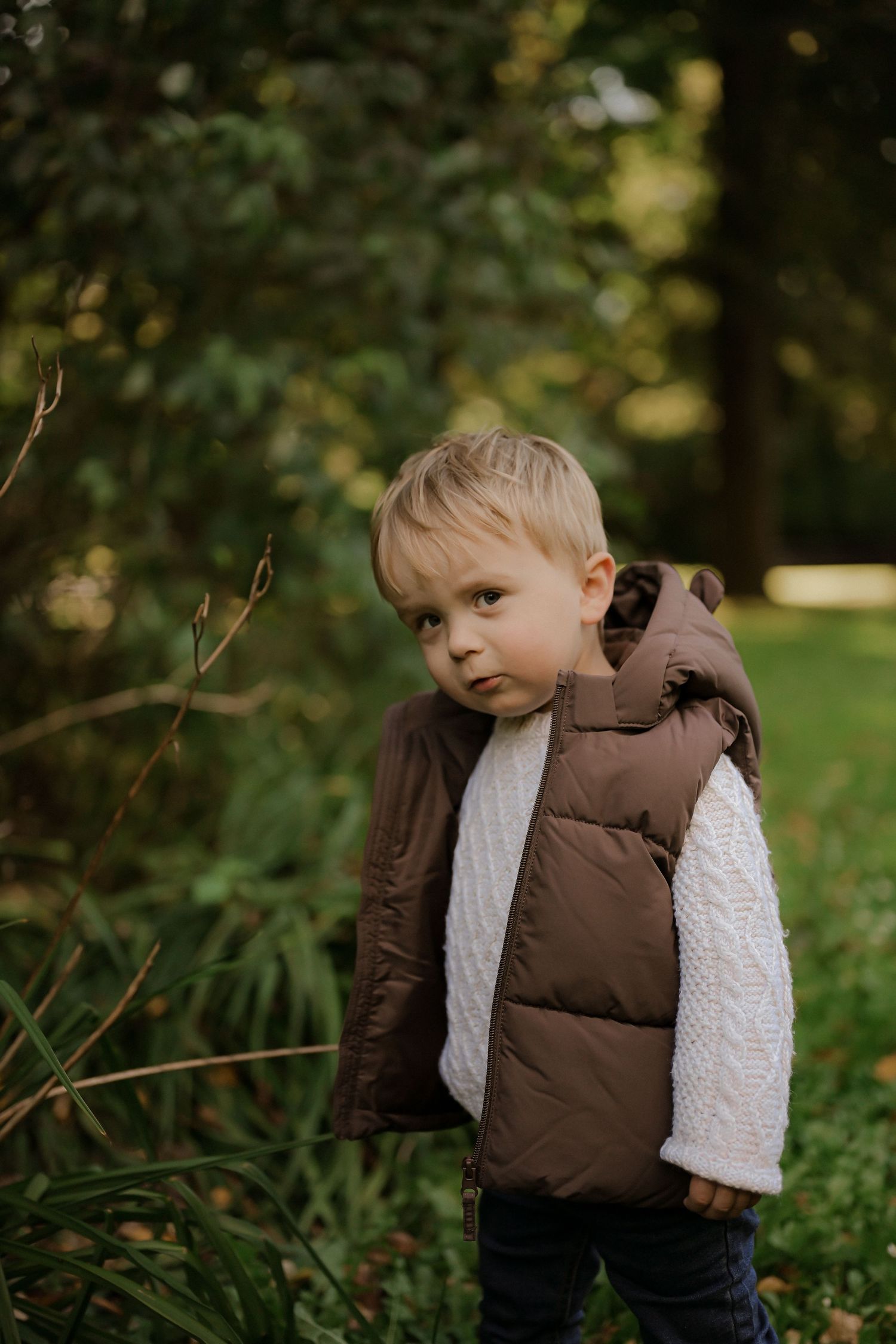 Young boy in brown puffer vest and white knit sweater standing outdoors among green trees in autumn park.