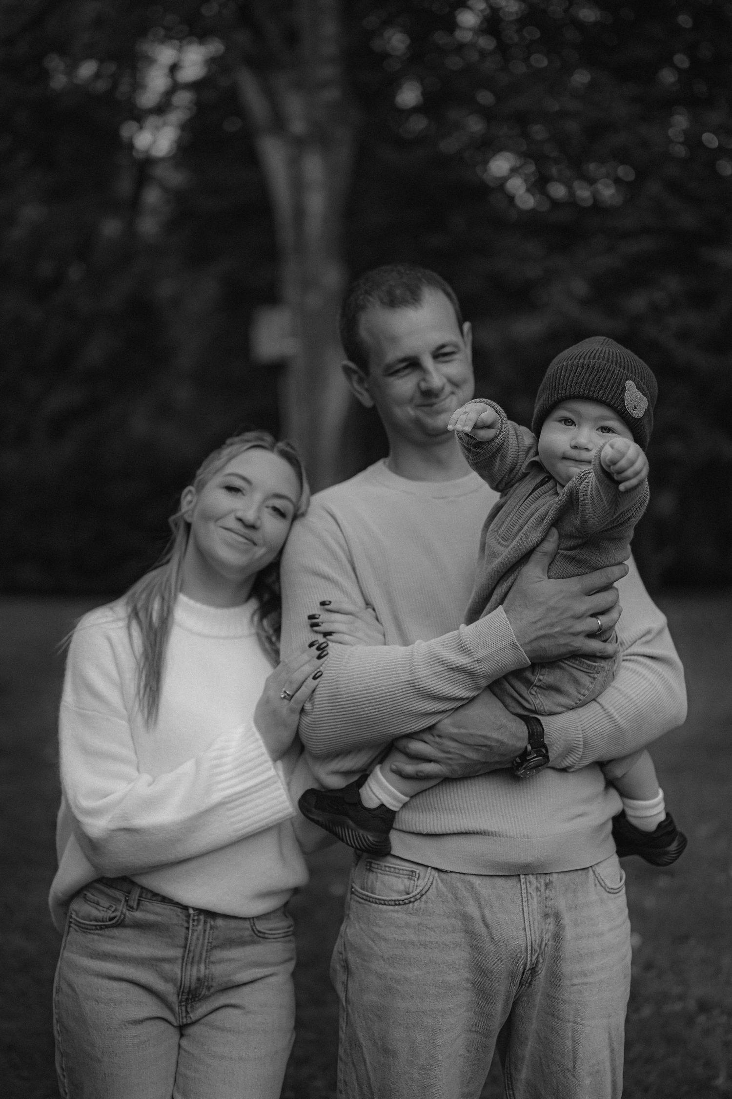Black and white family portrait of parents with baby in park setting.