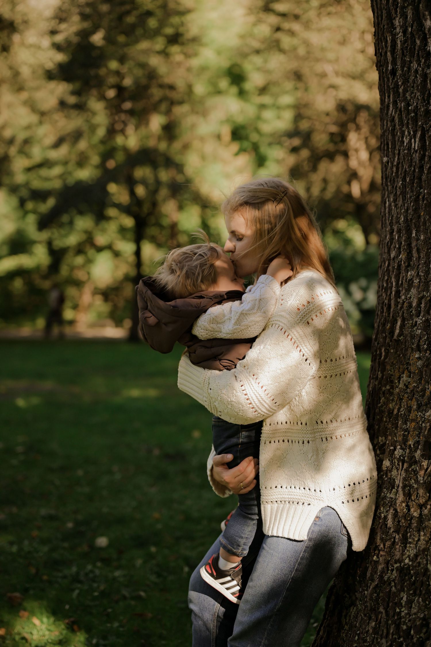 A mother tenderly kisses her young child while leaning against a tree in a lush green park.