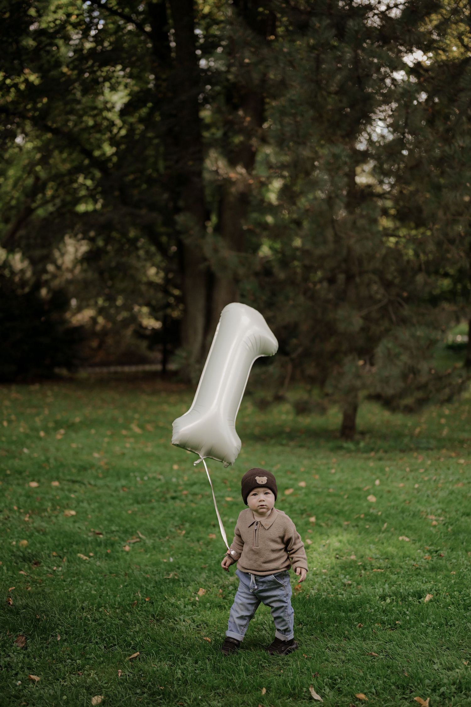 Toddler holding a number one balloon on green grass in a park, celebrating their first birthday outdoors.
