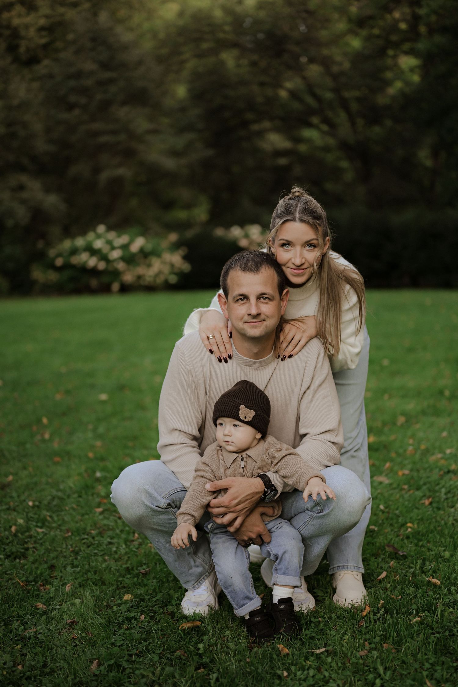 A happy young family with a baby boy poses outdoors on green grass with lush trees in the background.