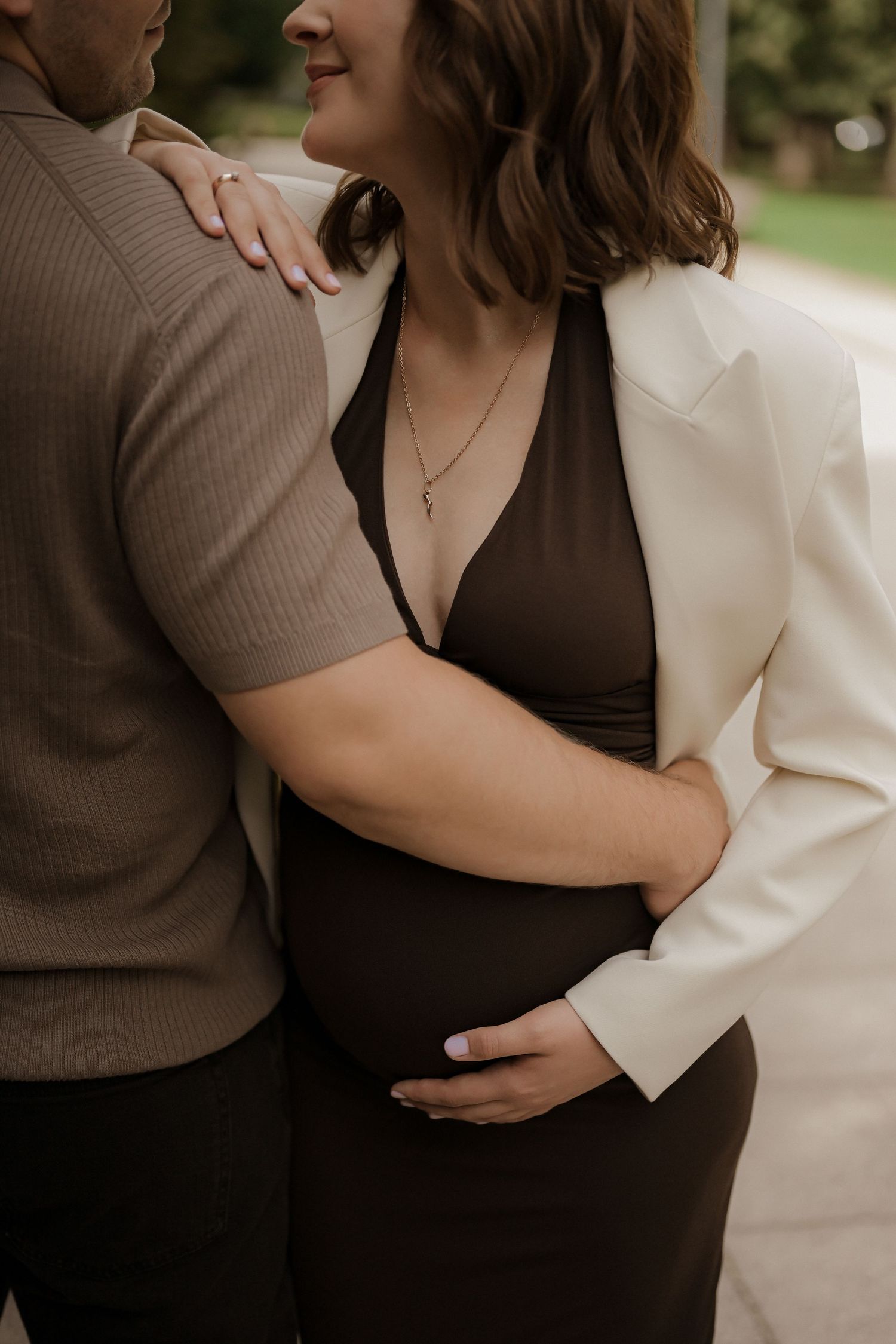 Pregnant woman in brown dress and white blazer cradles baby bump while partner embraces her outdoors.