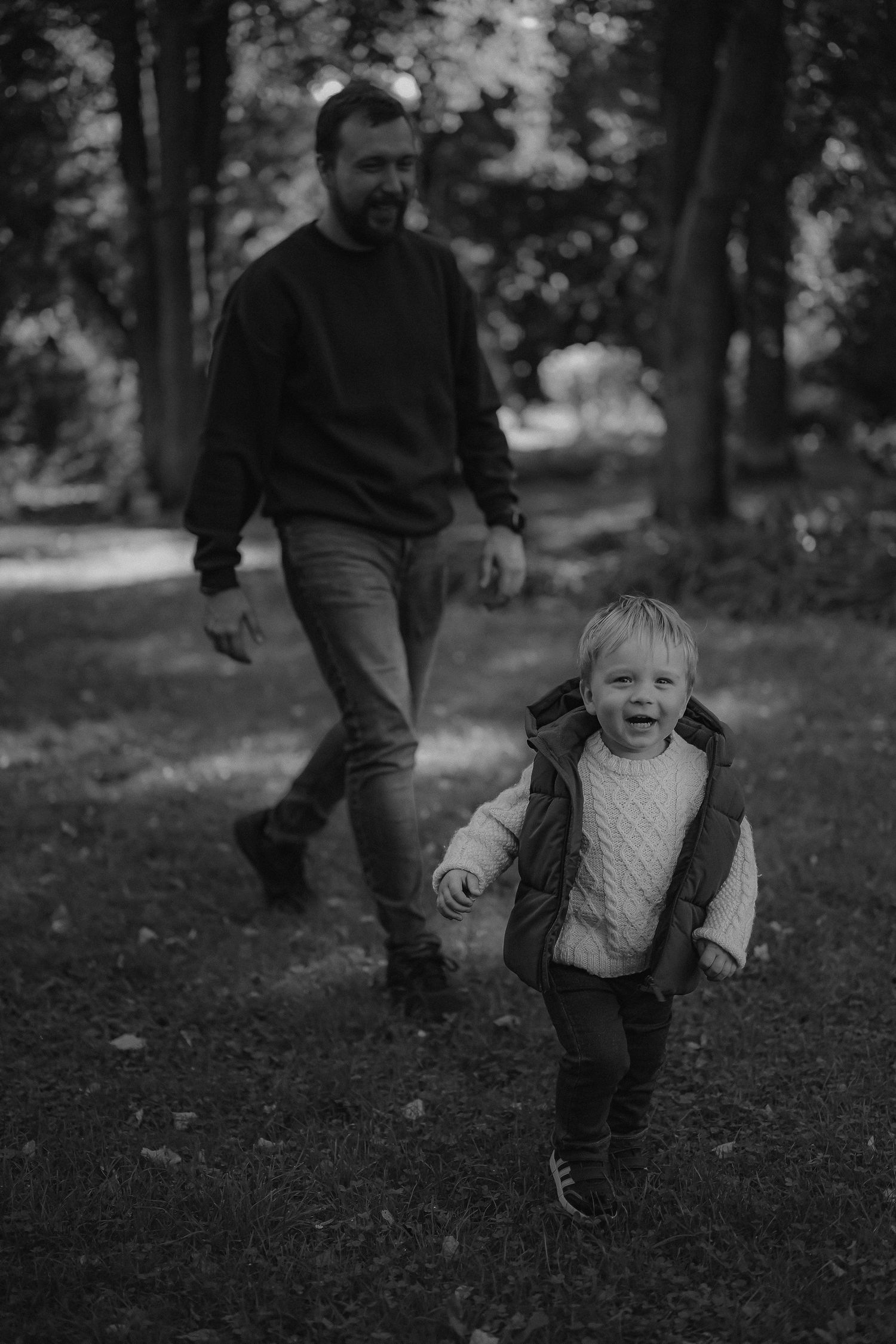 A black and white photo of a bearded father walking behind his laughing toddler in a park.