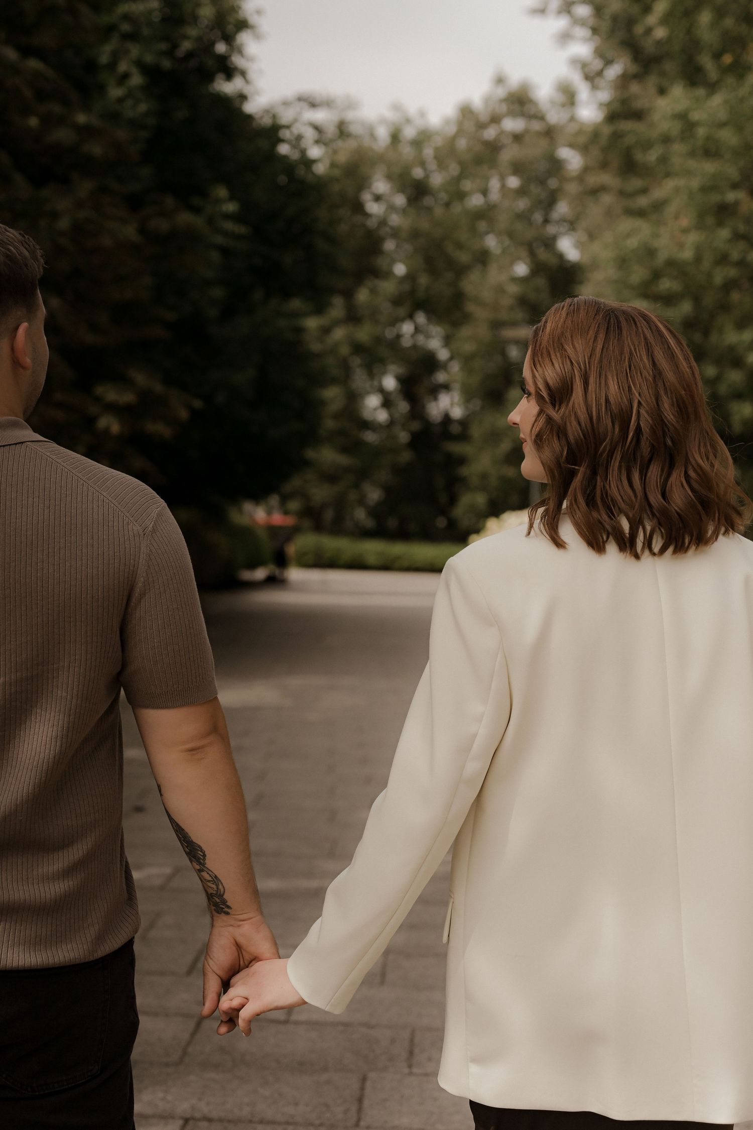 Couple holding hands while walking along a tree-lined park path, woman in white blazer and man in brown shirt with tattoo.