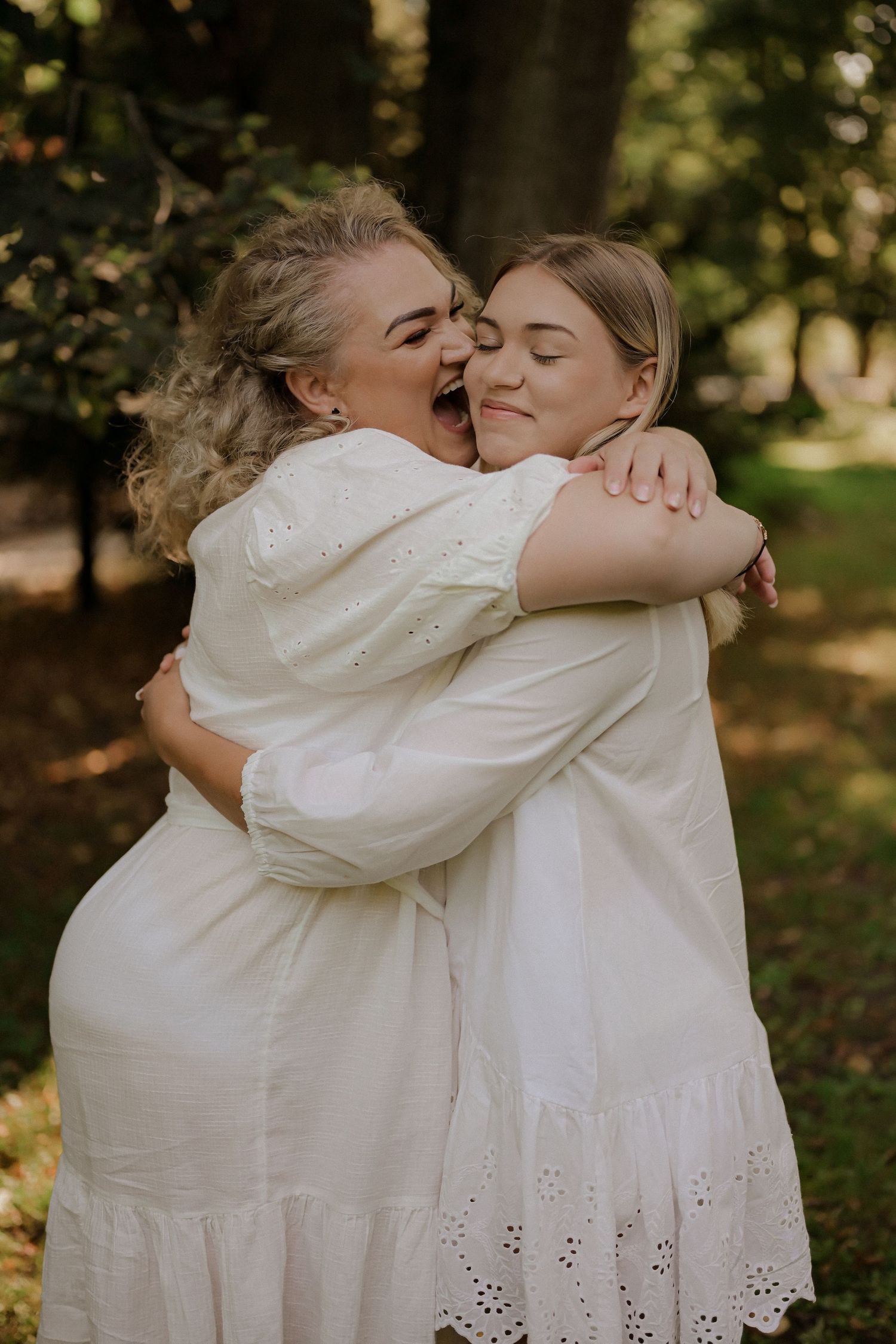 Two women in matching white dresses share a joyful hug and laugh outdoors.