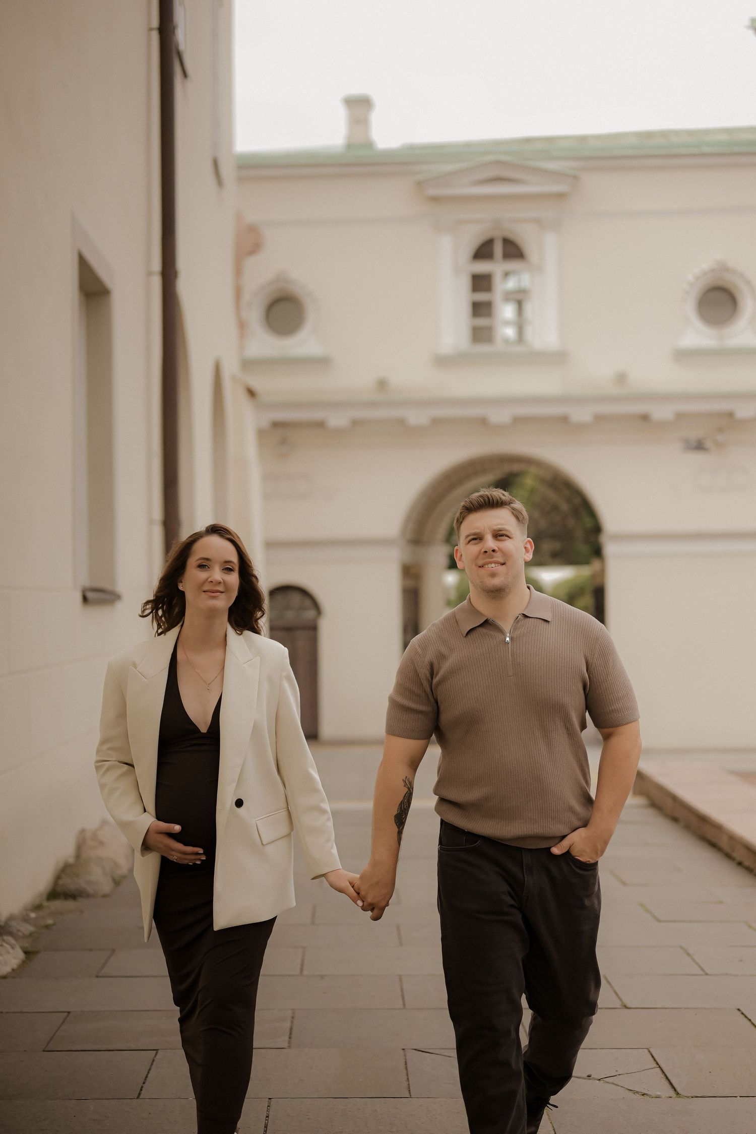 Pregnant woman in white blazer and man in brown polo hold hands walking past a classical European building.