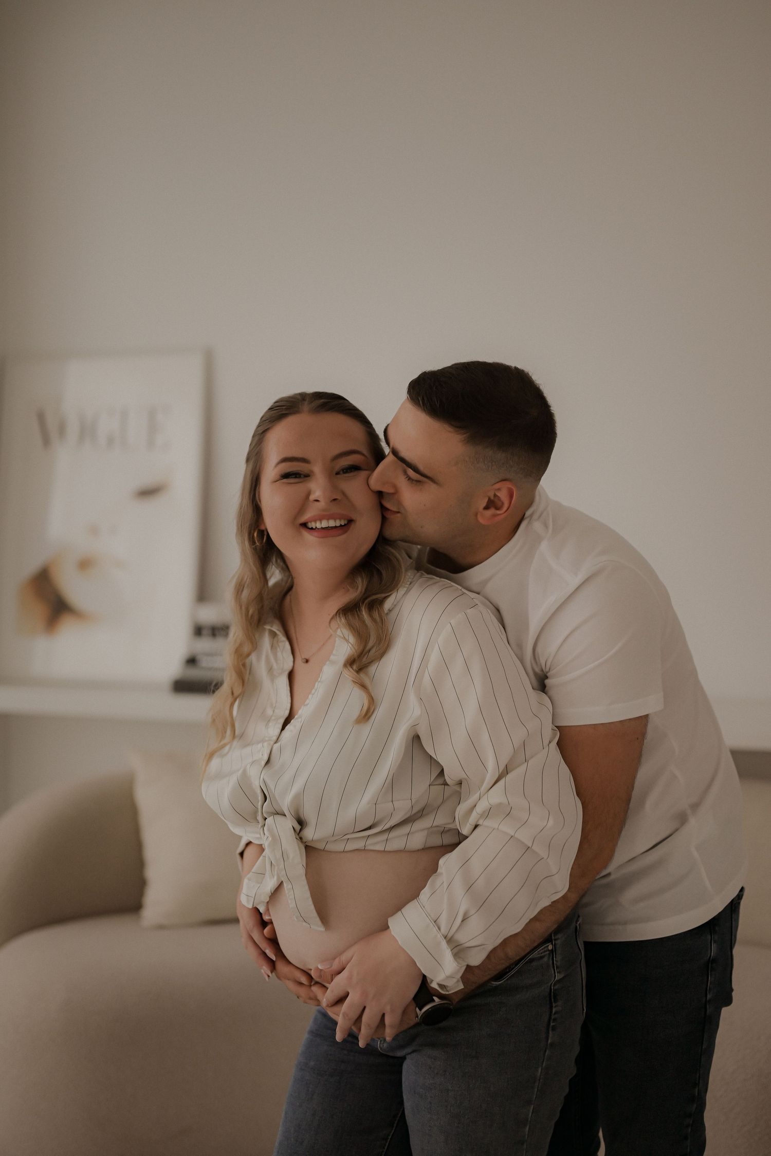 A couple shares a tender moment, man kissing woman's cheek while cradling her baby bump in a cozy white room.