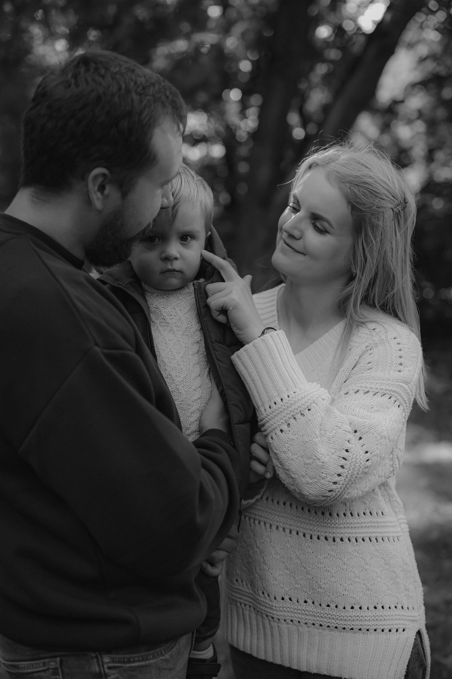 Black and white family portrait of parents lovingly looking at their toddler outdoors among trees.