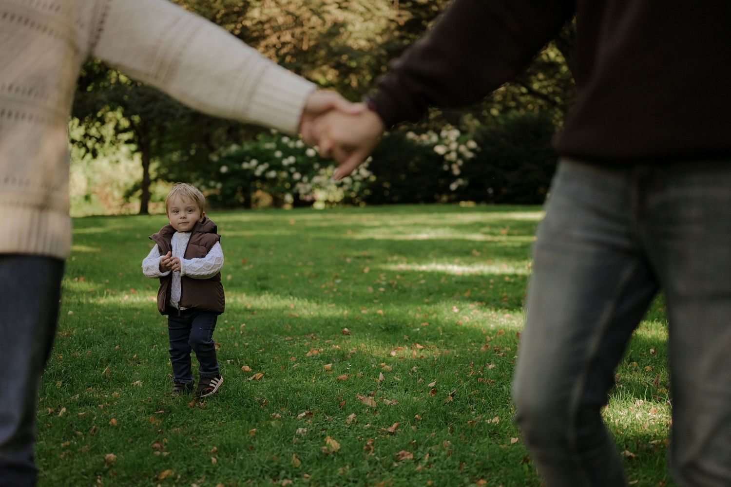 Toddler boy walking toward parents holding hands in a lush green park during autumn family photo session.