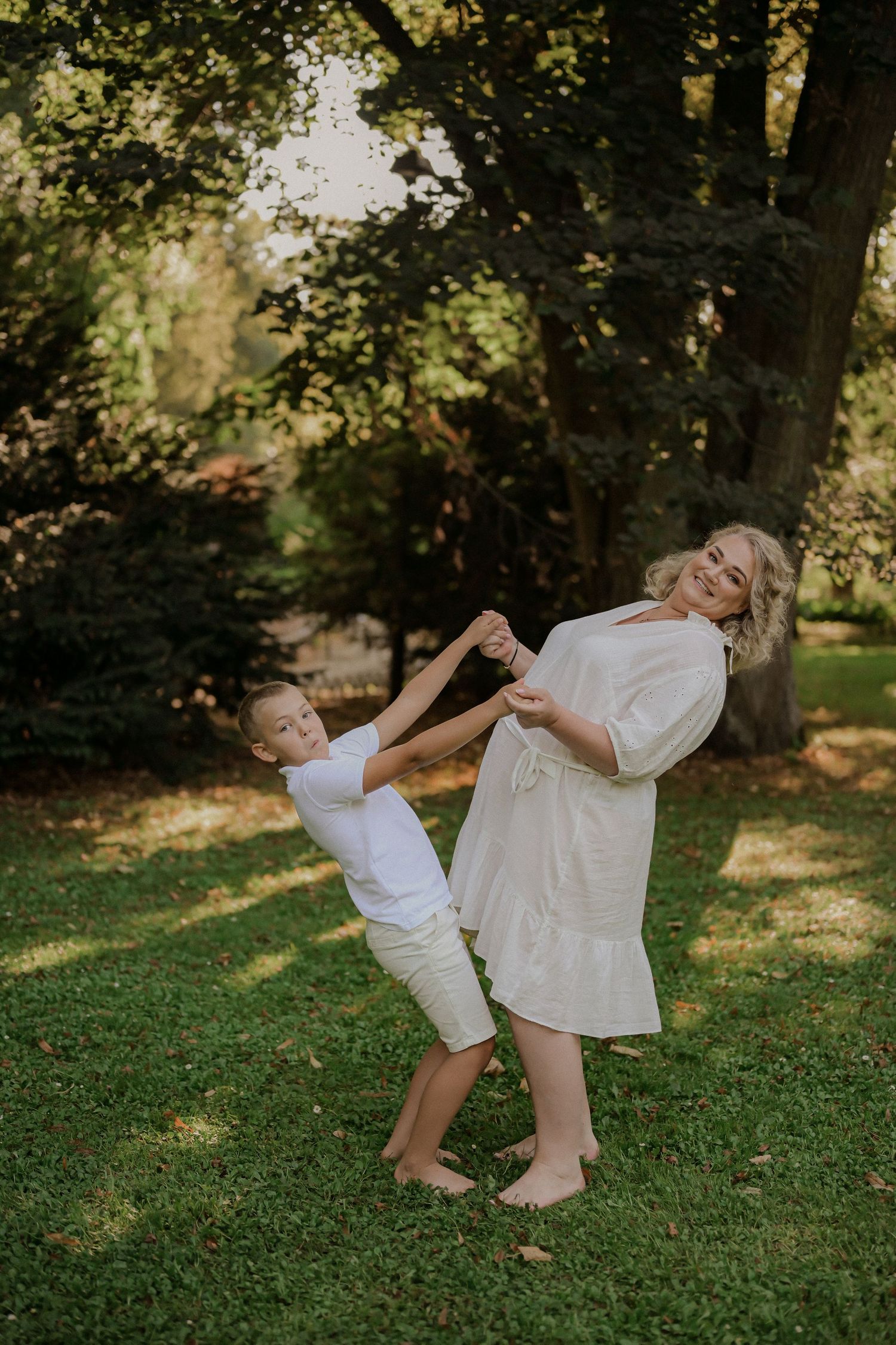 A woman in a white dress dances playfully with a young boy in white on green grass surrounded by trees.