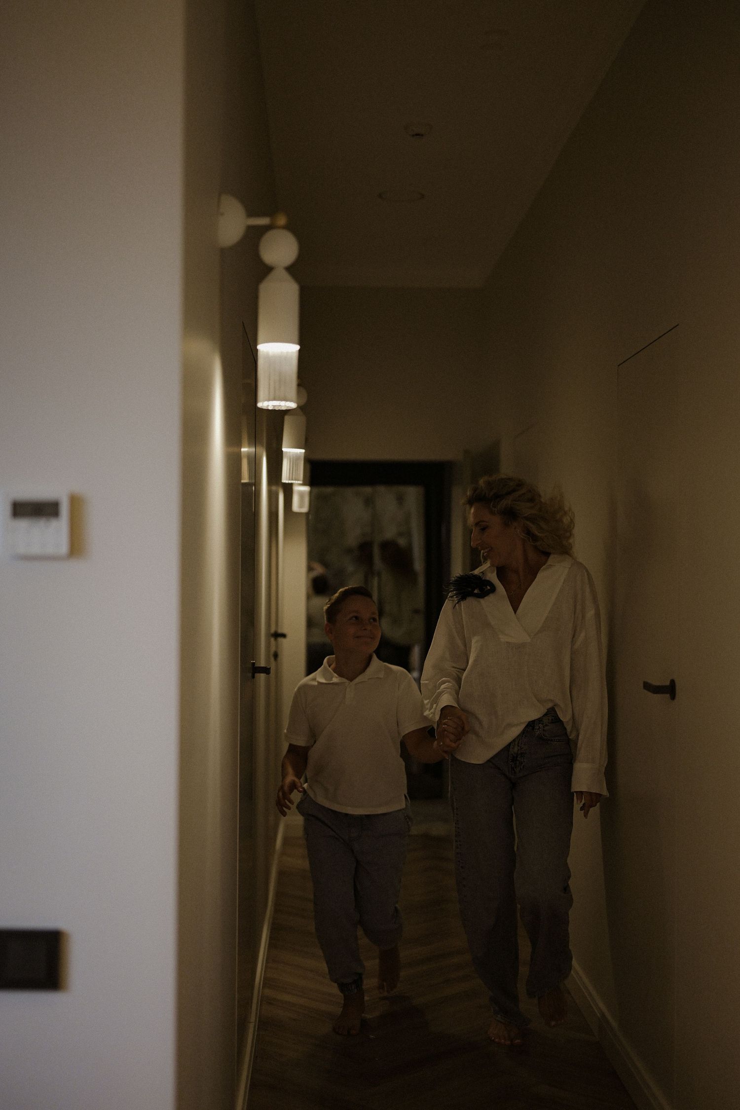 A woman and young boy walk hand in hand down a warmly lit modern hallway together.
