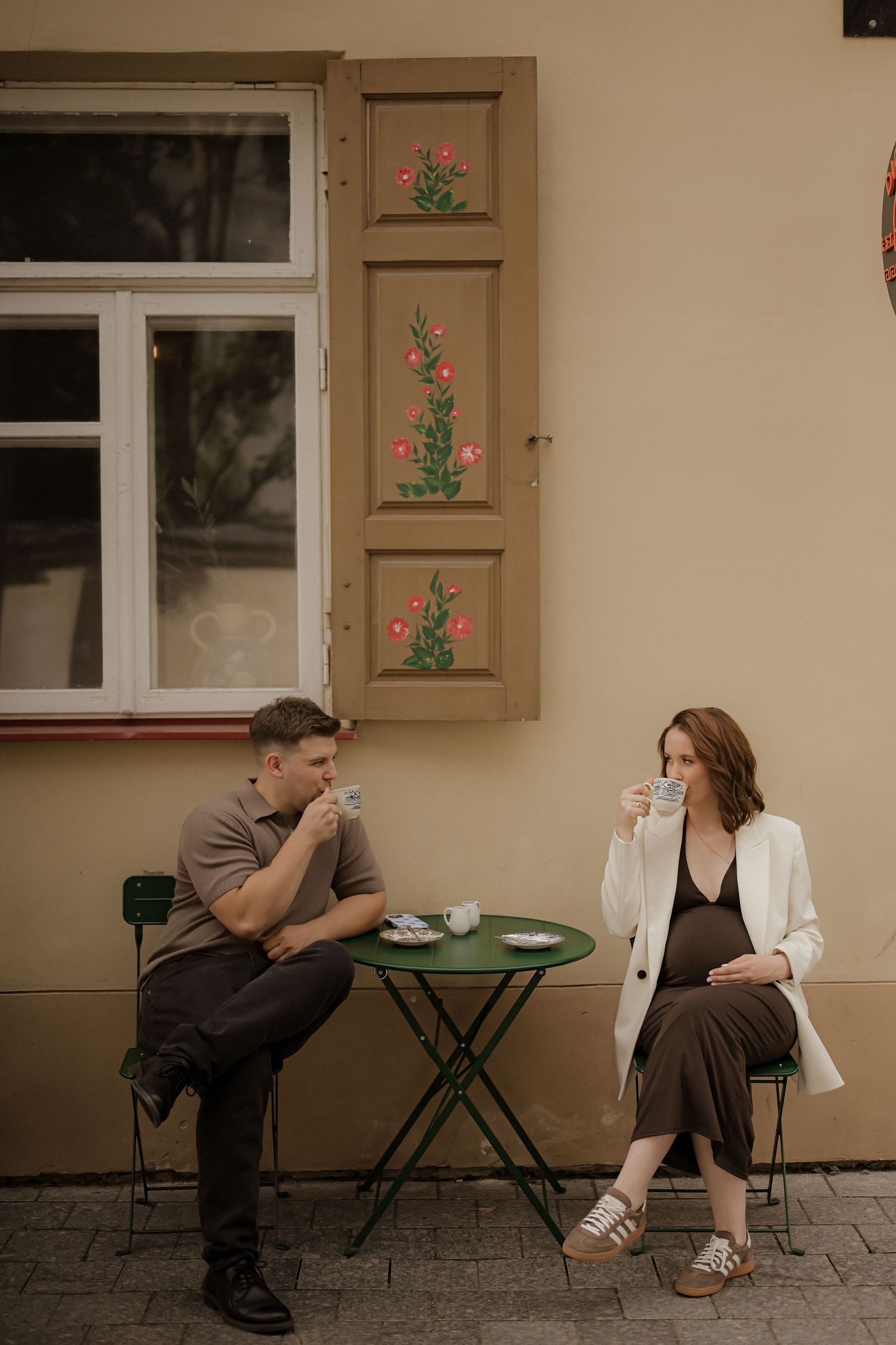 A couple enjoys coffee at a charming outdoor cafe table beside a decorated European building wall.