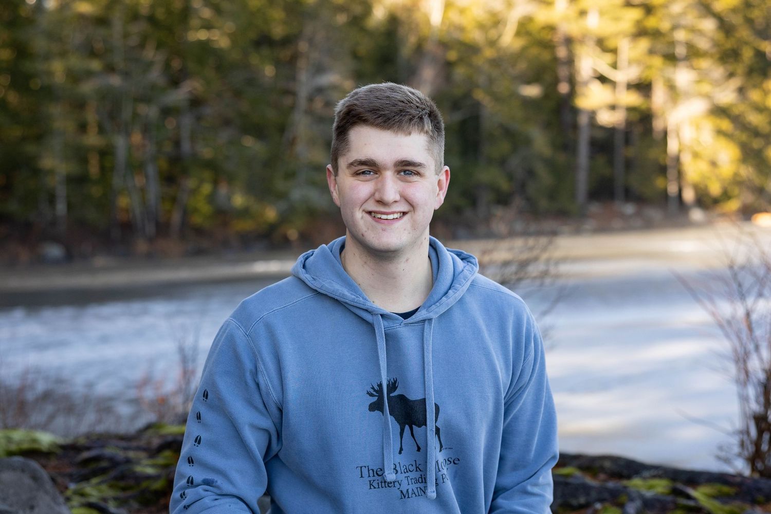 Smiling young man in a blue hoodie poses outdoors near a frozen lake in winter.