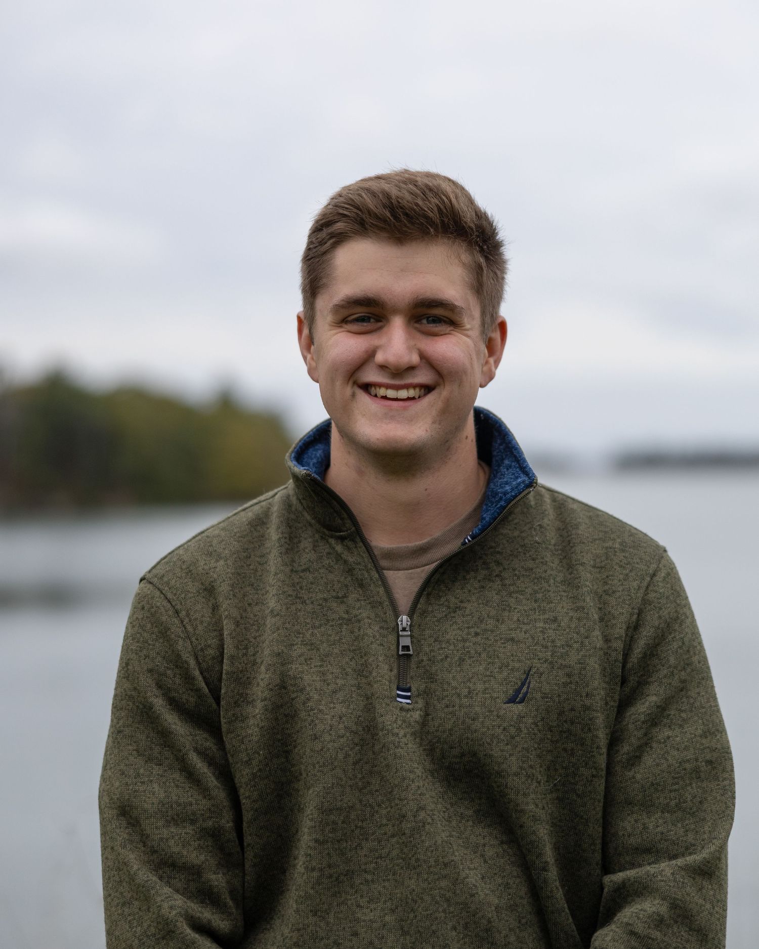 Smiling young man in a green Nautica pullover stands outdoors near a lake on a cloudy day.