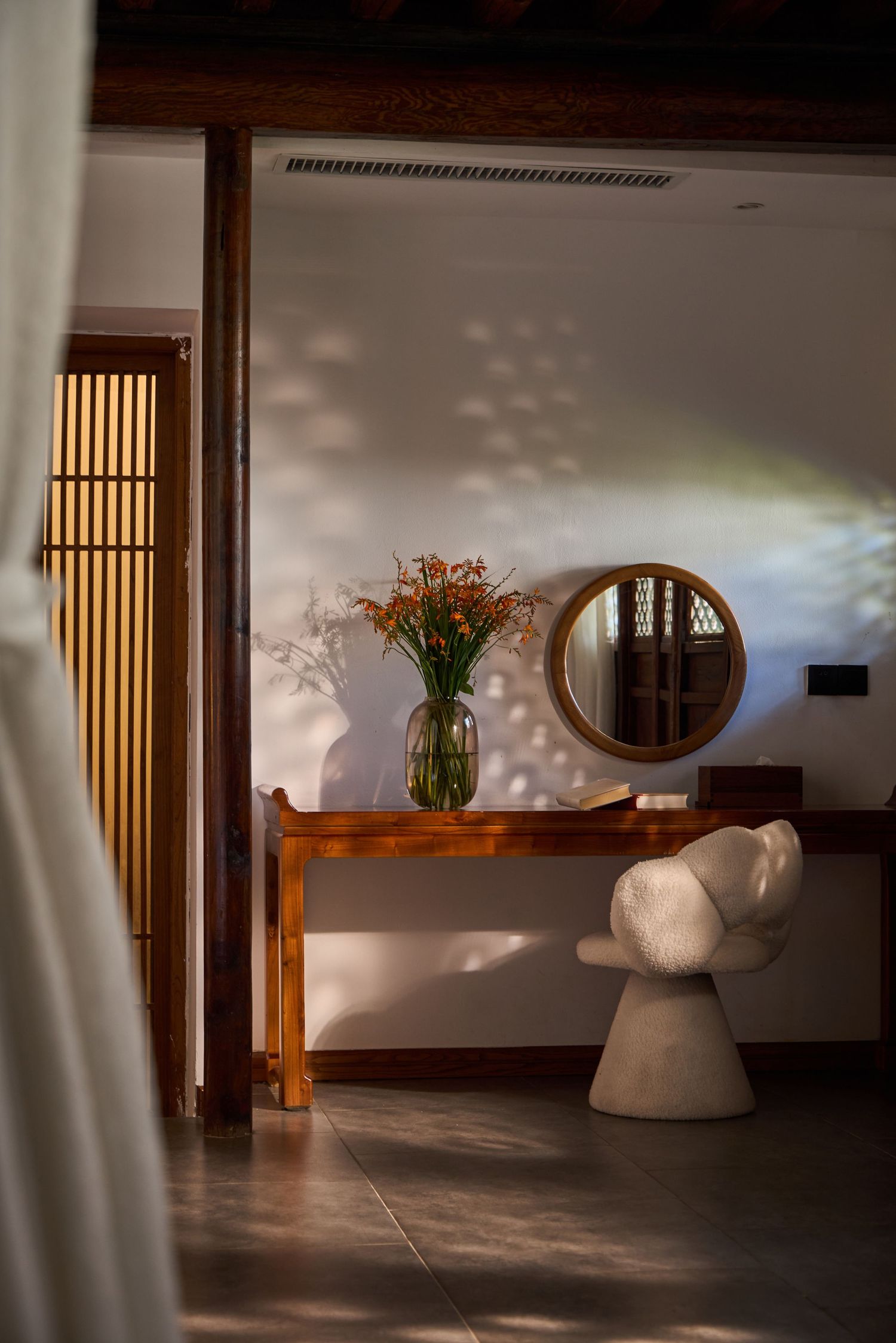 Elegant entryway with wooden console table, vase of orange flowers, round mirror and sculptural white chair in soft sunlight.