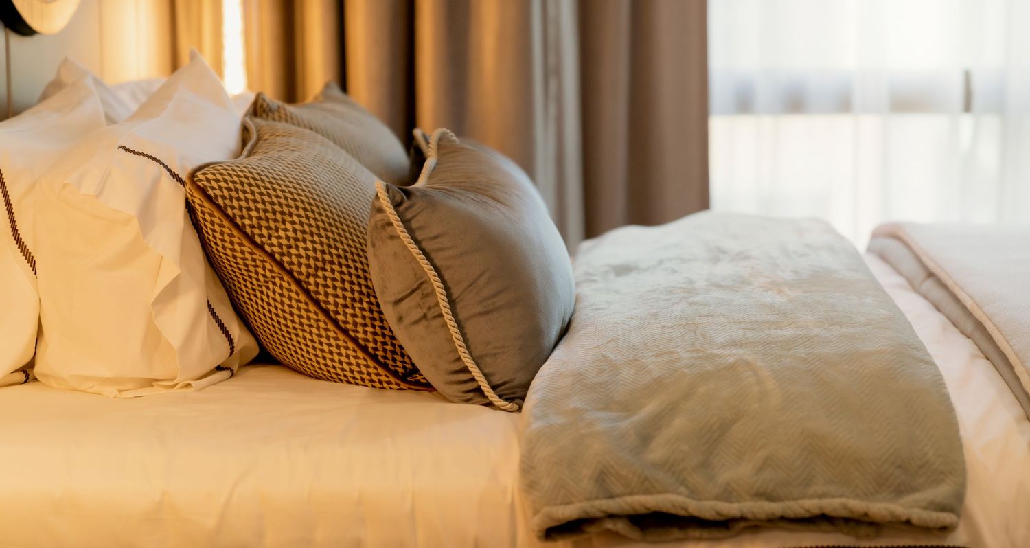 Close-up of a neatly made hotel bed with beige and patterned pillows, a gray folded throw blanket and soft cream curtains nearby.