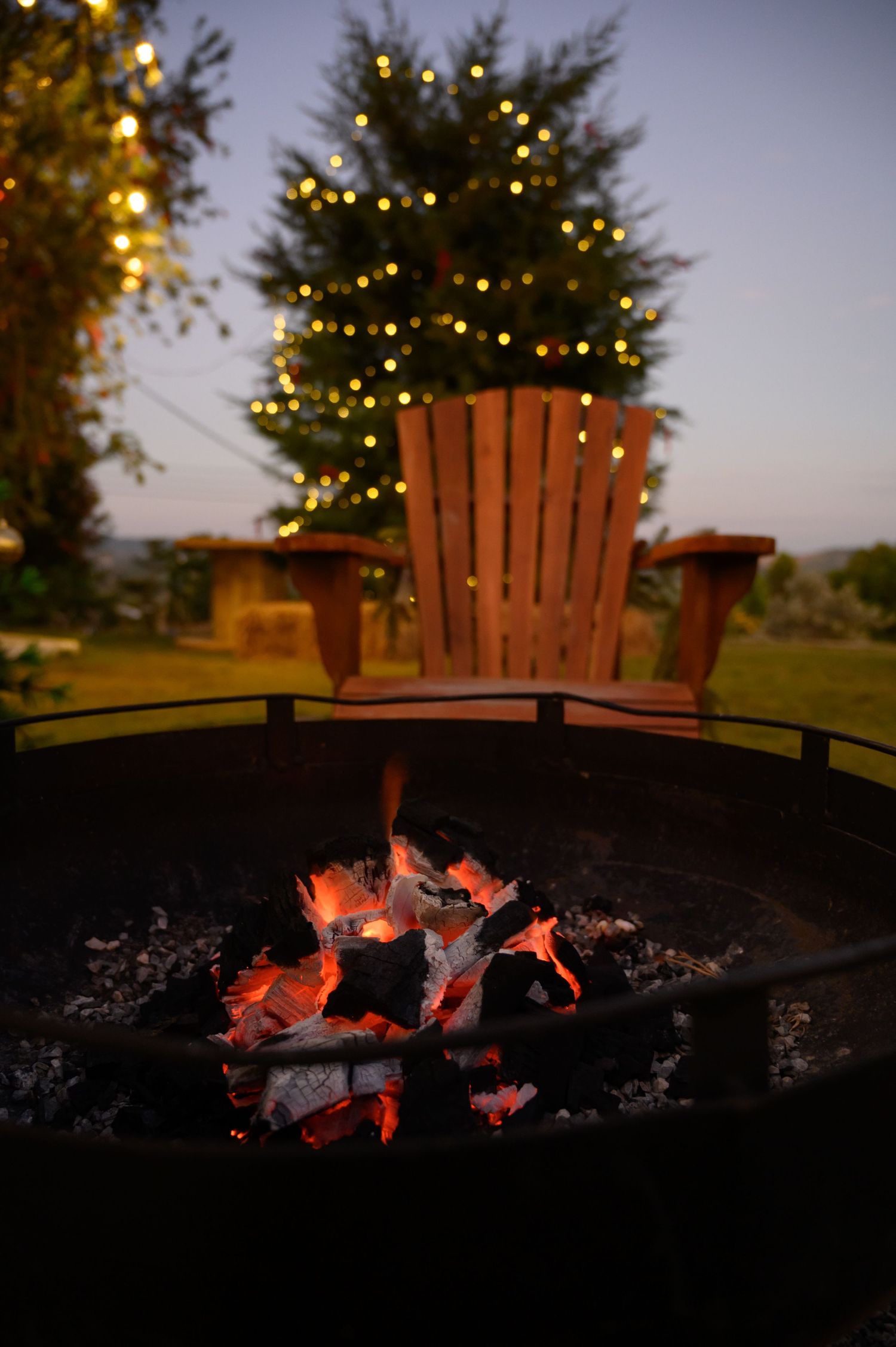 Glowing fire pit at dusk with a wooden Adirondack chair and a Christmas tree strung with warm lights behind.