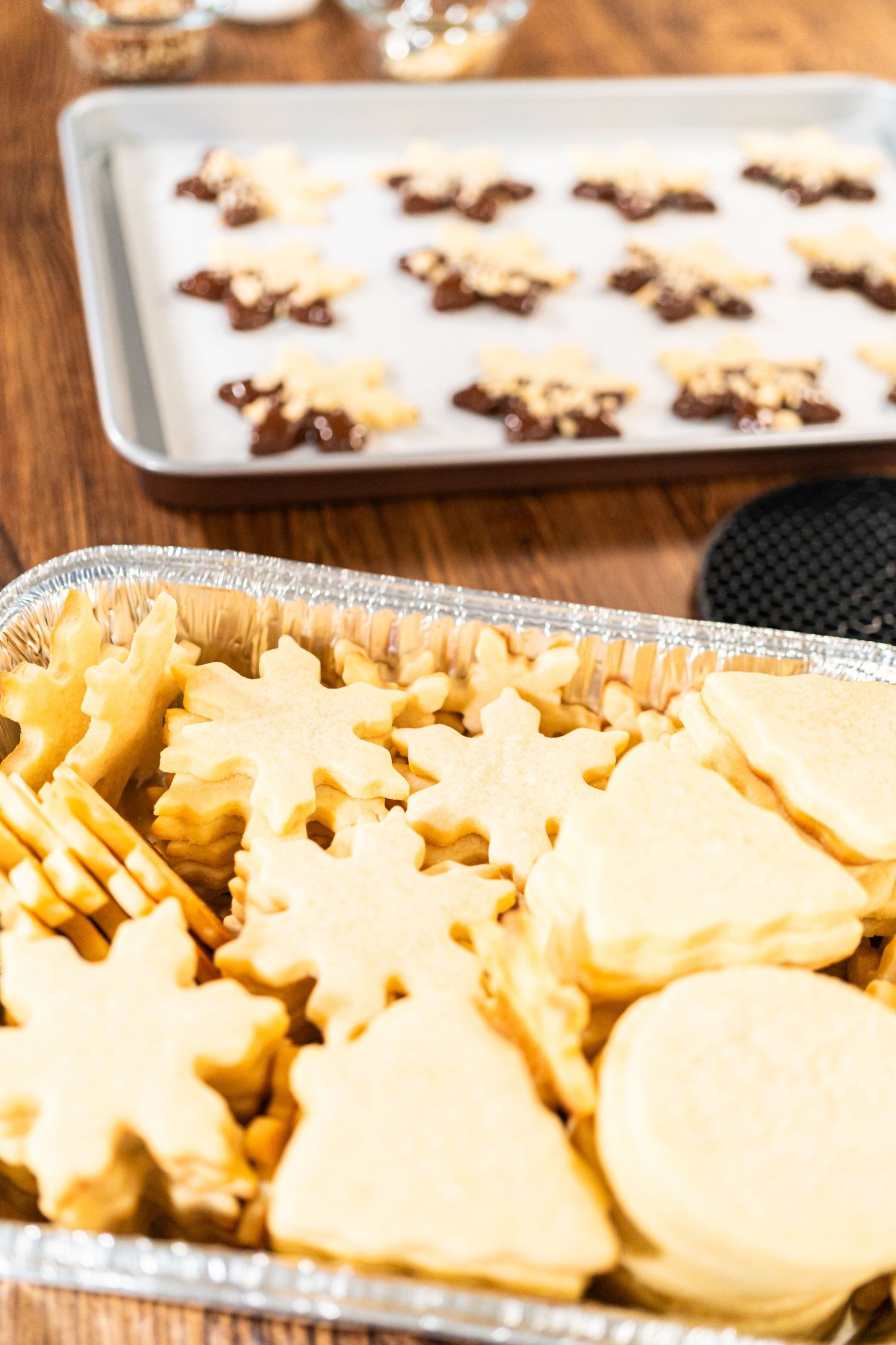 Star and tree shaped sugar cookies in a foil pan, with chocolate-dipped decorated cookies on a baking tray behind.