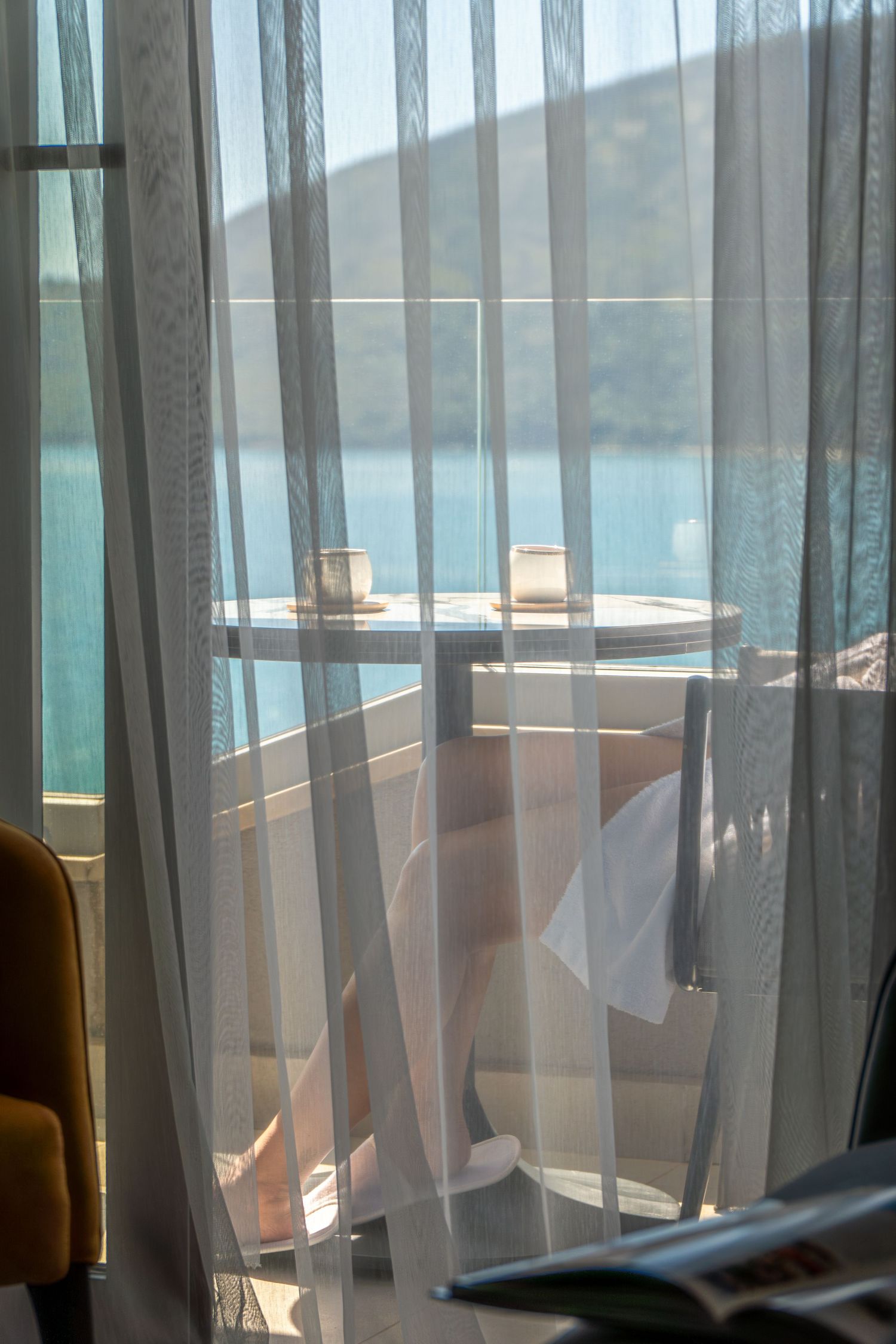 Sheer curtains frame a balcony table with two coffee cups overlooking a calm turquoise bay and hillside.