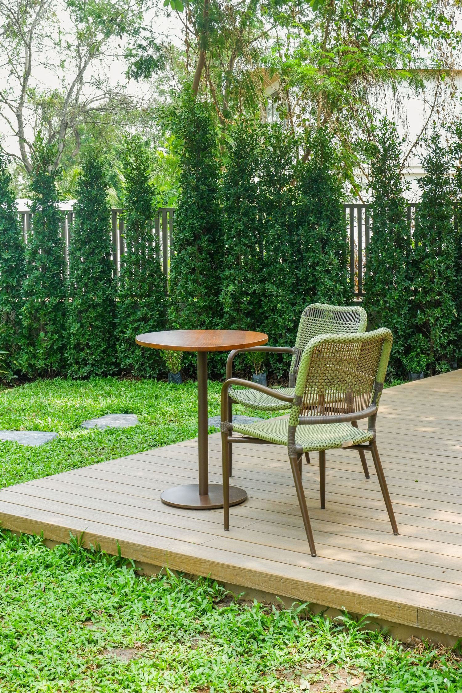 Round wooden bistro table and two green woven chairs on a wooden garden deck beside a lawn and tall green hedge.
