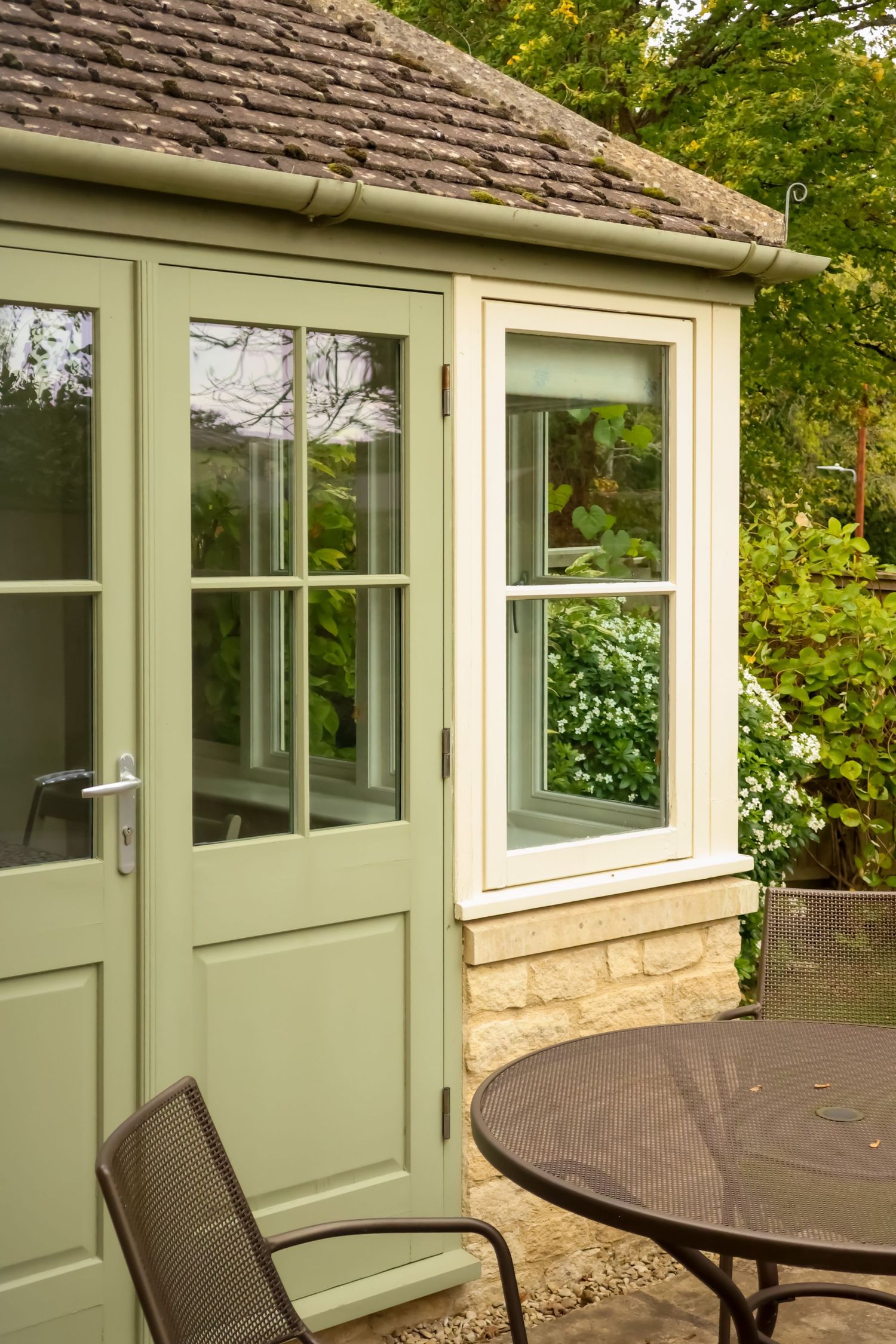 Sage green French doors and a sash window on a Cotswold stone cottage overlooking a garden patio with a metal table and chairs.