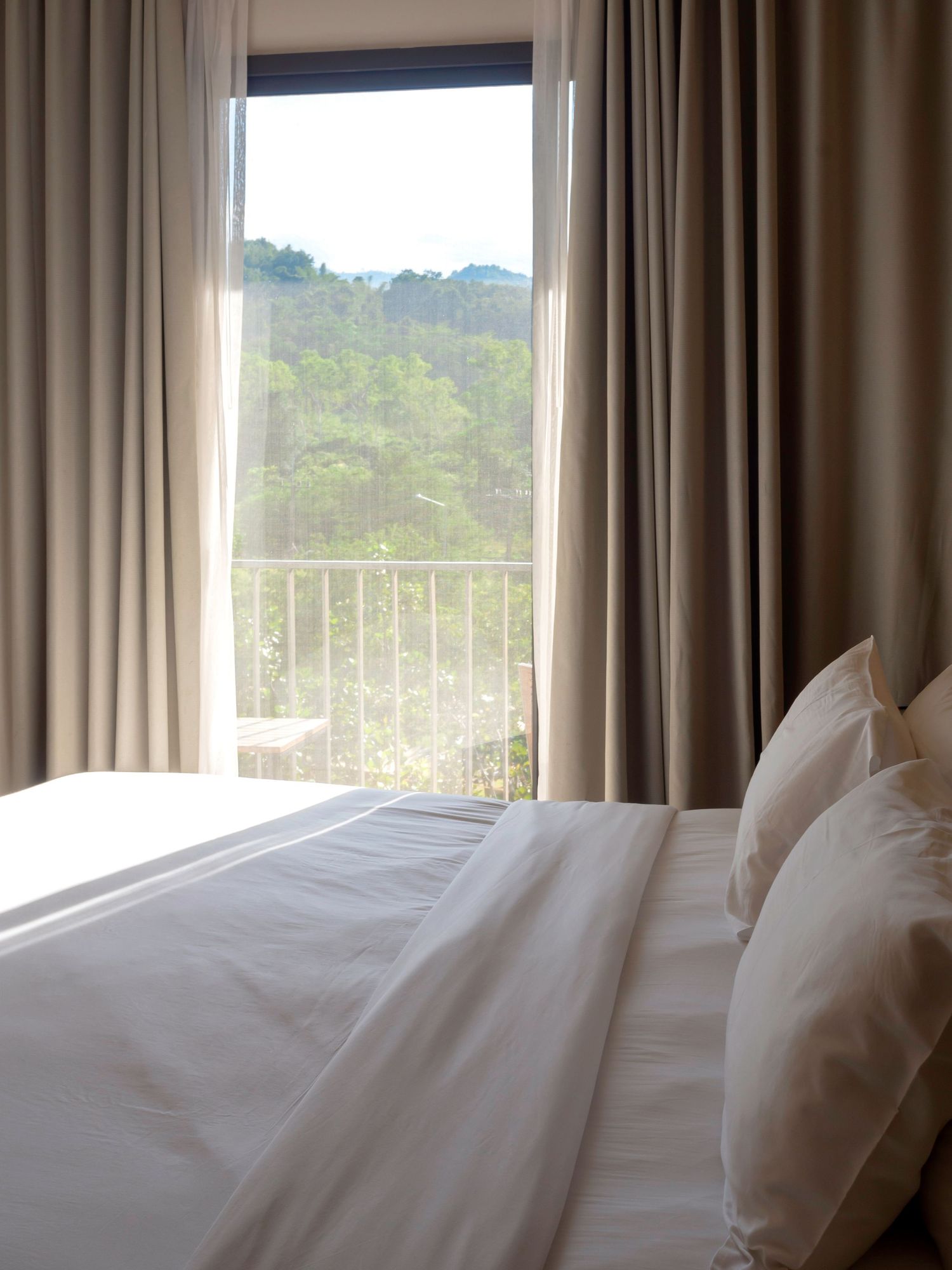 Bed with white linens and pillows beside floor-to-ceiling windows with taupe curtains overlooking a lush green hillside.