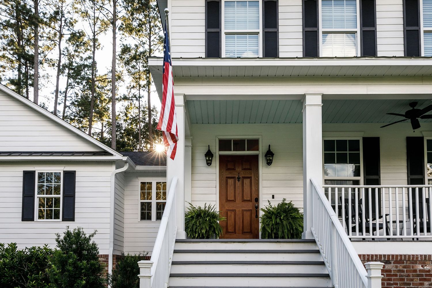 Front porch of a white house with a wooden door, black shutters, an American flag and steps lined with shrubs.