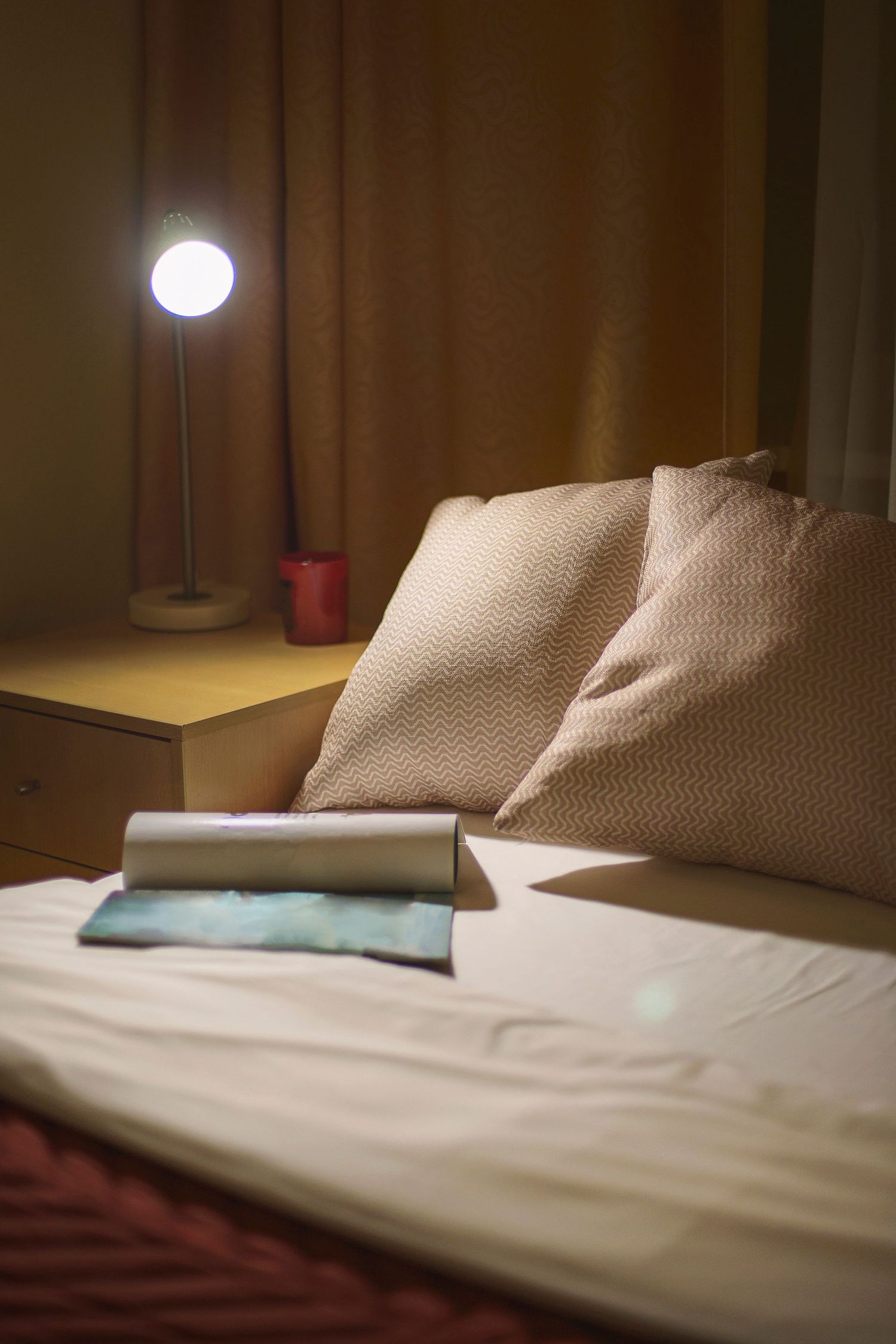 Bed with white sheets and beige pillows, an open magazine and a glowing bedside lamp on a wooden nightstand in a softly lit bedroom.