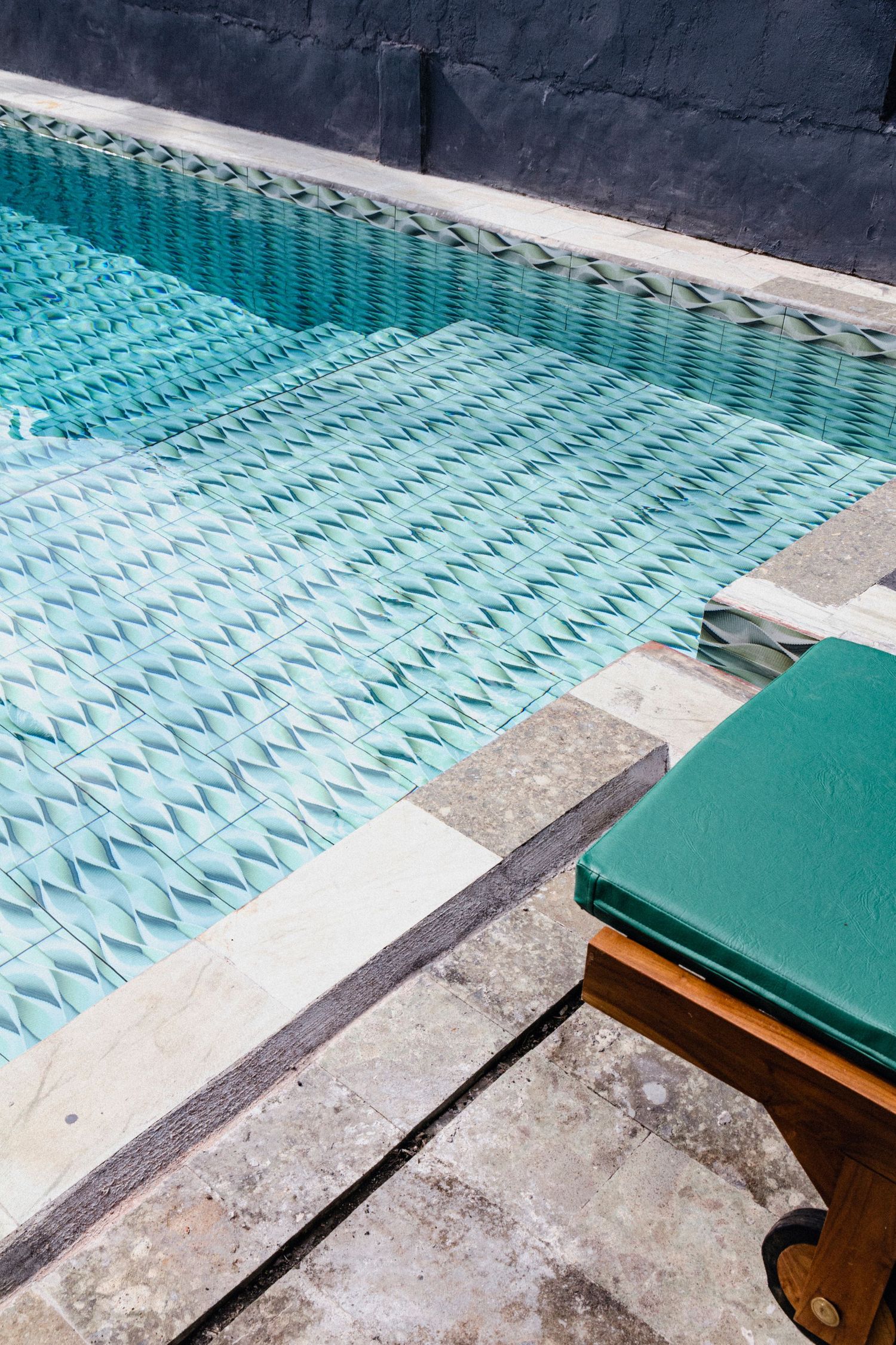 Turquoise patterned pool tiles with a stone edge and a green cushioned wooden sun lounger beside the water.