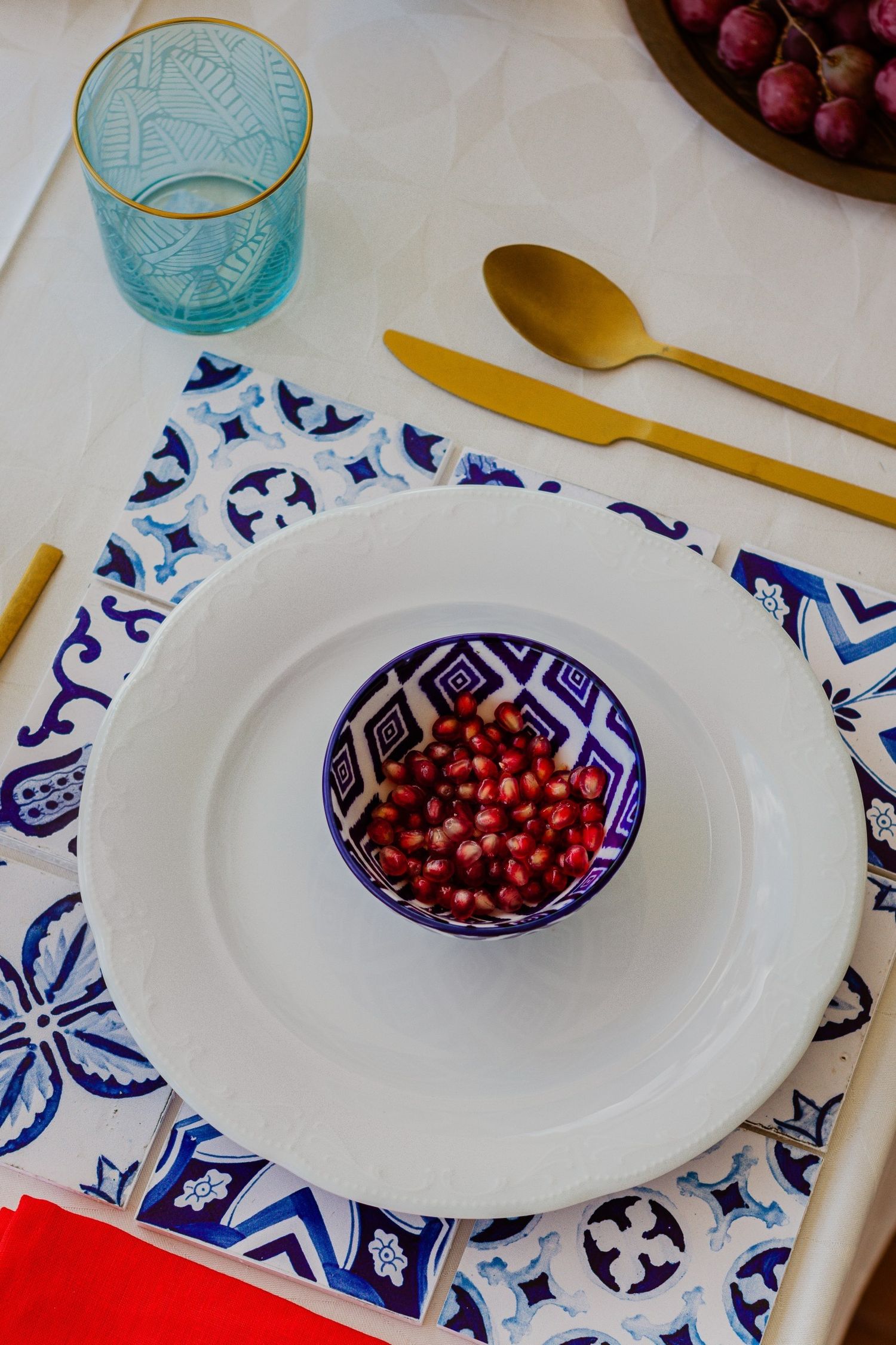 Elegant table setting with pomegranate seeds in a blue patterned bowl, gold cutlery, tile placemat, and blue glass.