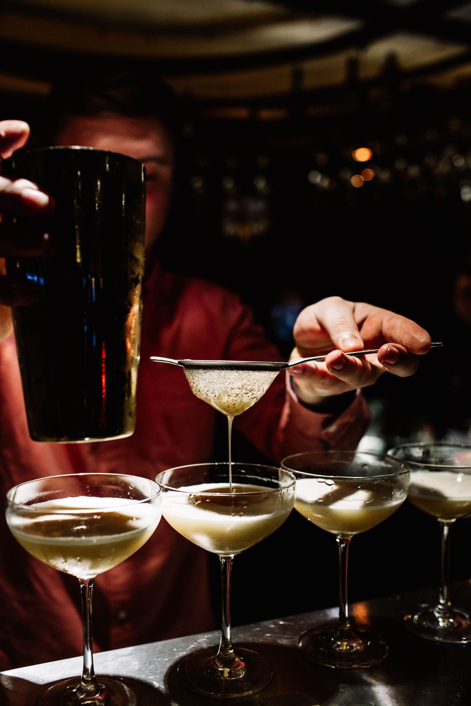 Bartender in a red shirt strains a cocktail through a fine strainer from a shaker into a row of coupe glasses at a dimly lit bar.