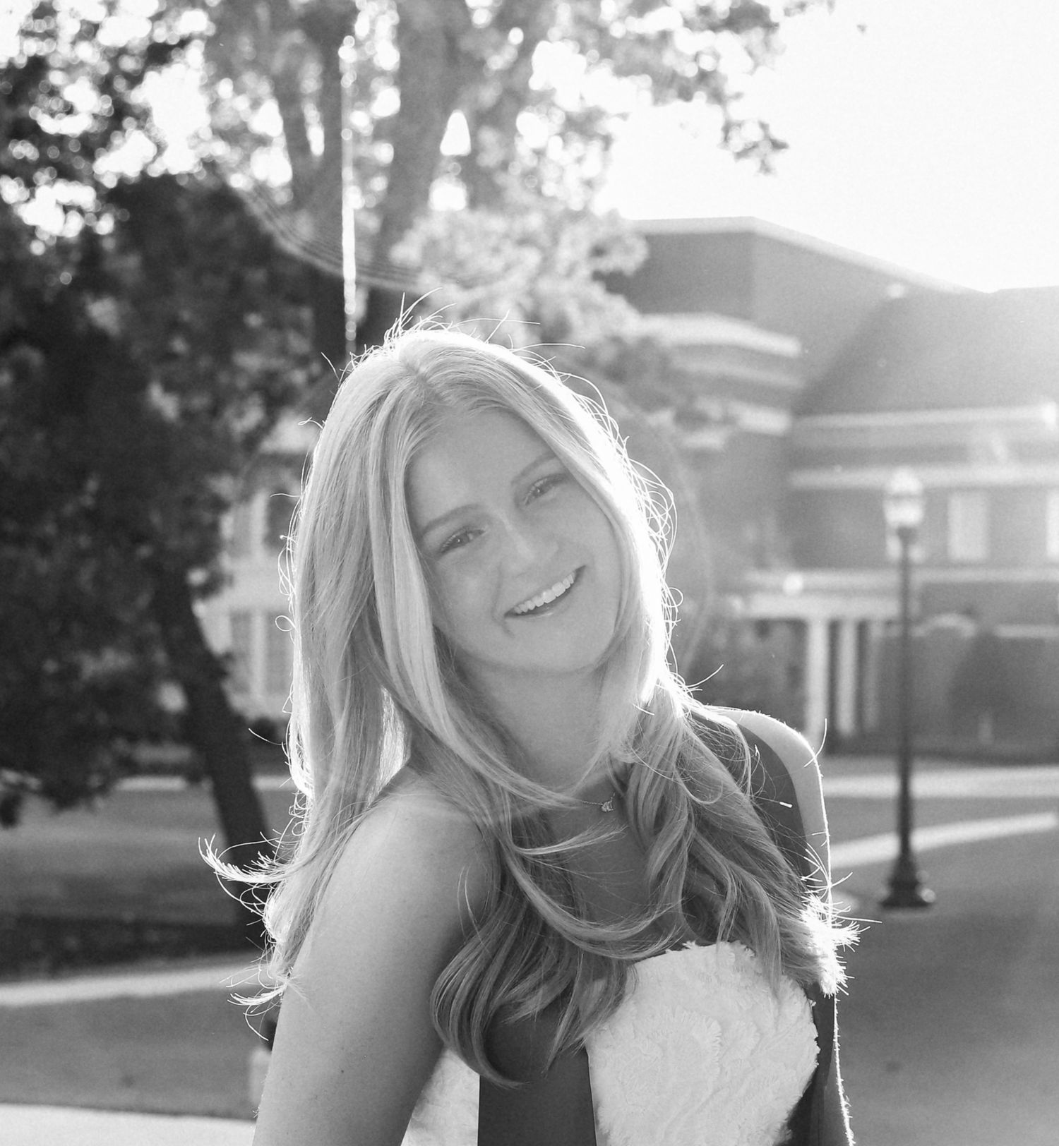 Smiling young woman with long wavy blonde hair poses outdoors in black and white with sunlit trees and a brick building behind her.