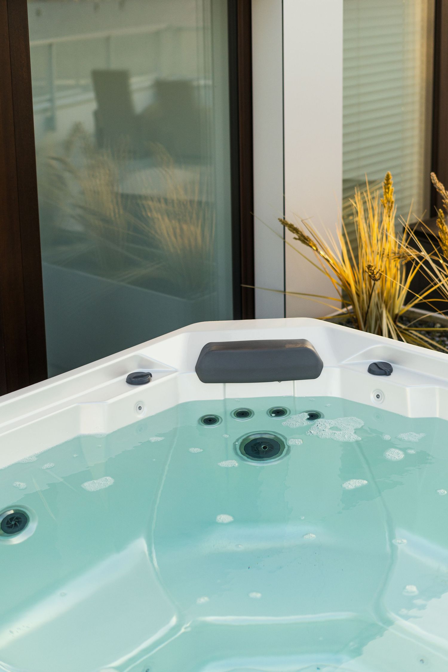 Close-up of a white outdoor hot tub with bubbling turquoise water, jets and a headrest beside tall ornamental grasses.