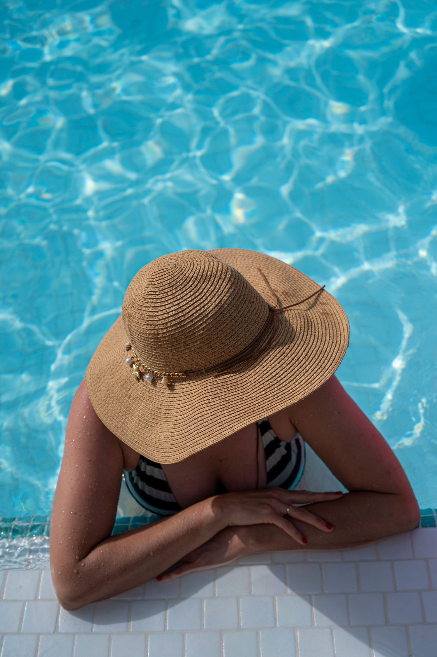 Woman in a straw sun hat and striped swimsuit rests her arms on the edge of a sparkling blue swimming pool.