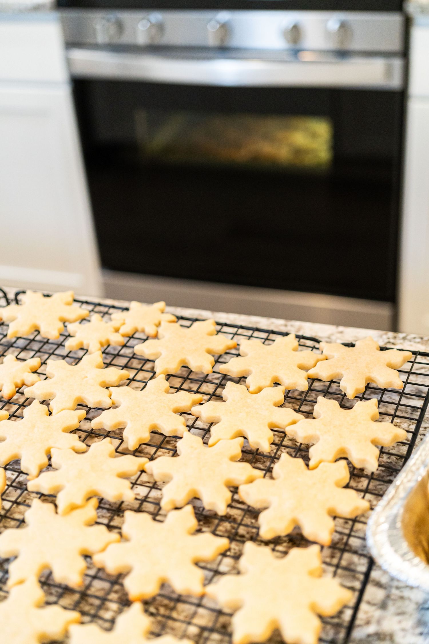 Freshly baked snowflake-shaped sugar cookies cooling on a wire rack on a granite counter in front of a kitchen oven.