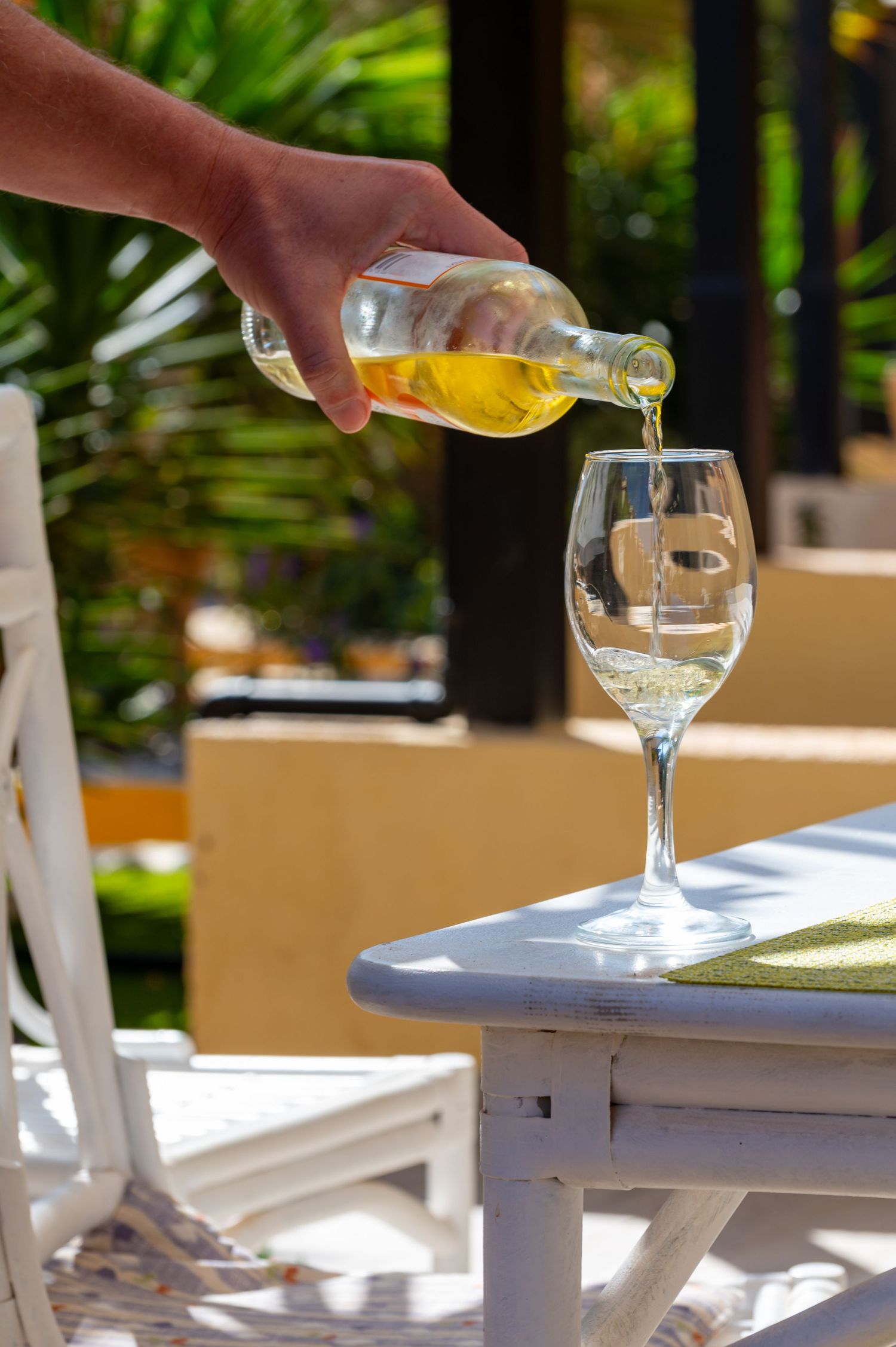 Hand pouring chilled white wine from a bottle into a stemmed glass on a white patio table in a sunny garden.