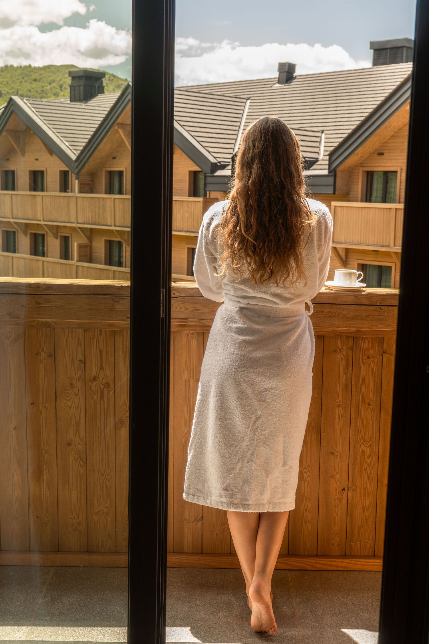 Woman in a white bathrobe stands barefoot on a wooden balcony with a cup of tea, overlooking timber chalets.