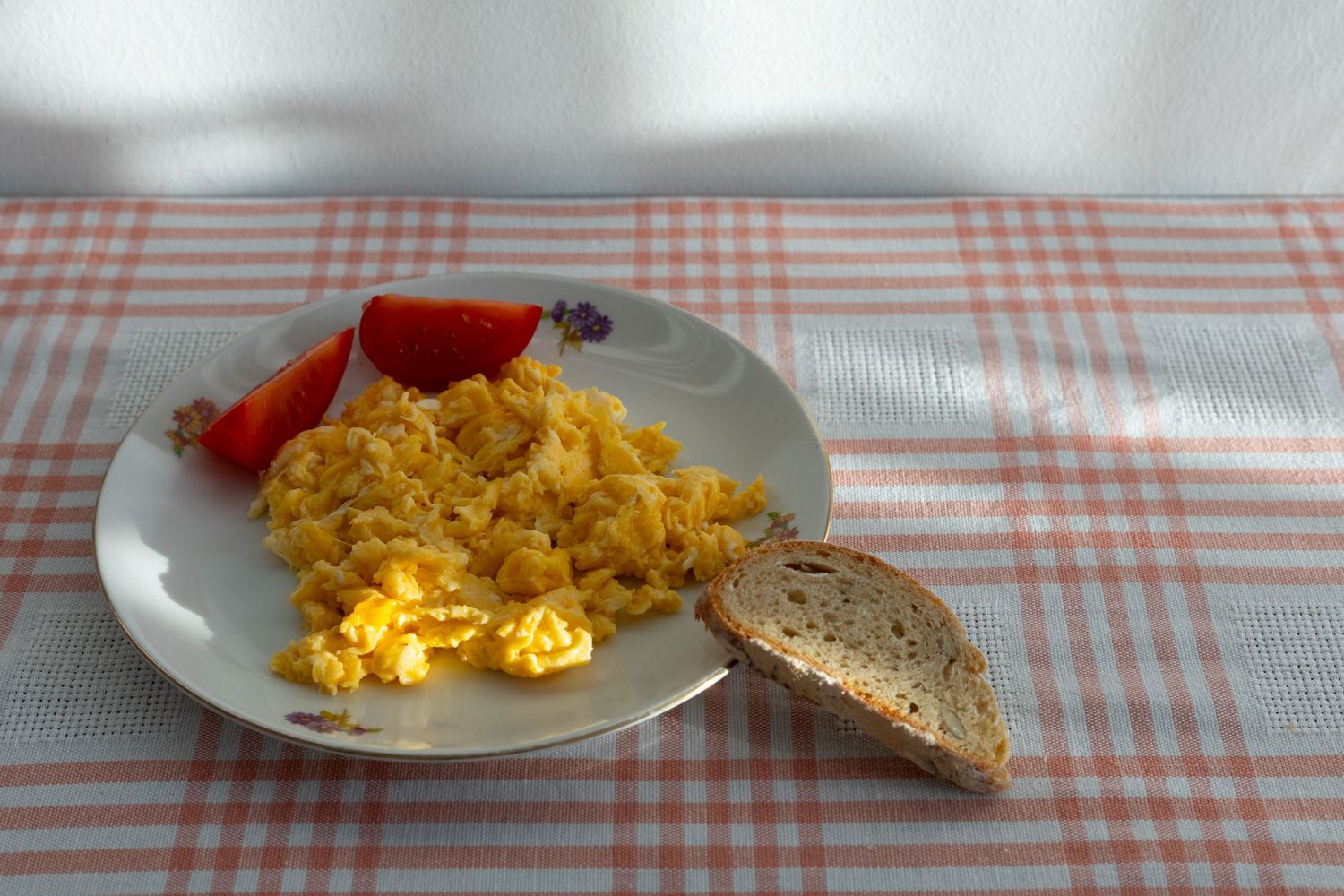 Plate of scrambled eggs with fresh tomato slices and a piece of bread on a red checkered tablecloth.