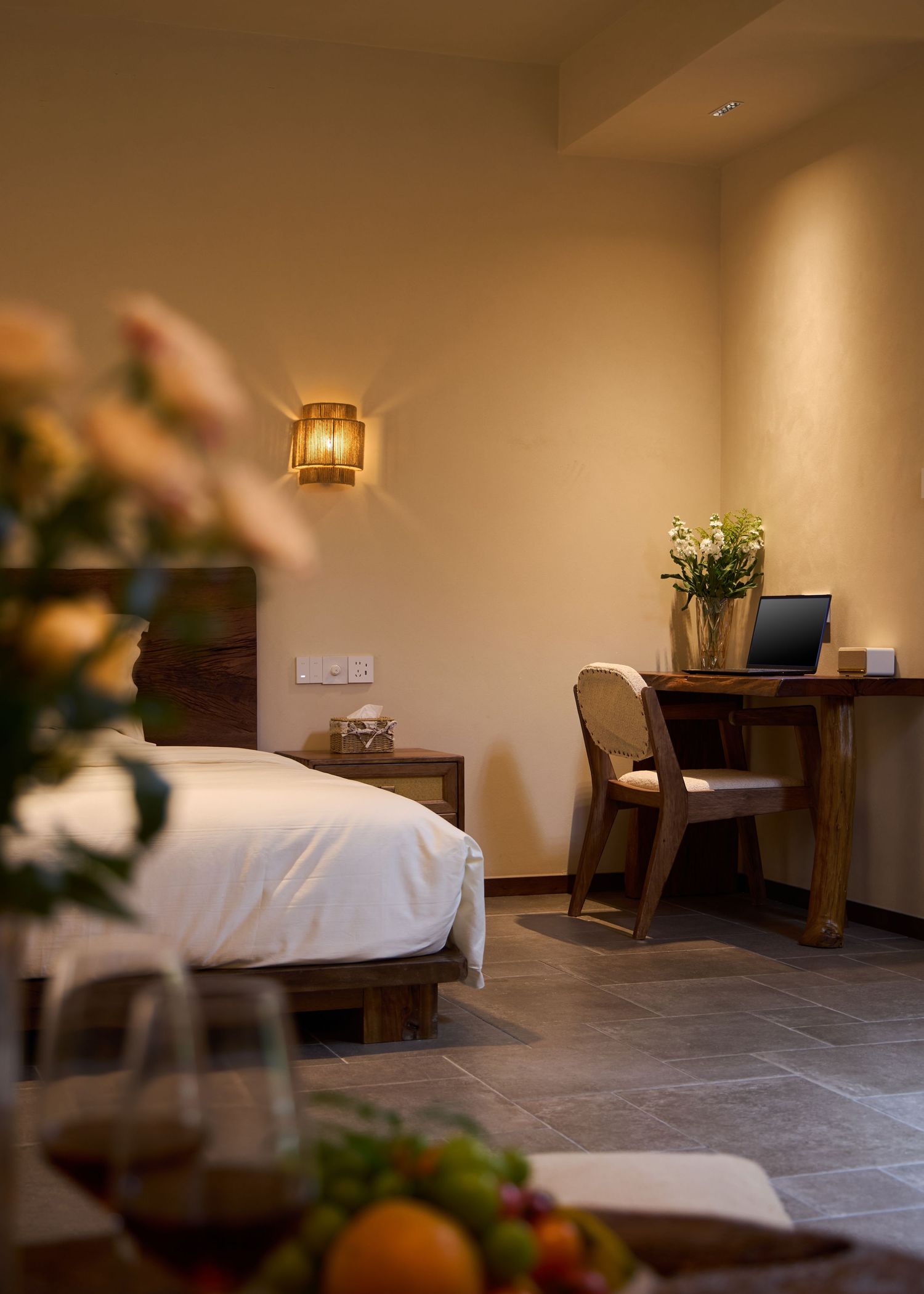 Warm hotel room with a white bed, glowing wall sconce, wooden desk with laptop and a fruit bowl in the foreground.