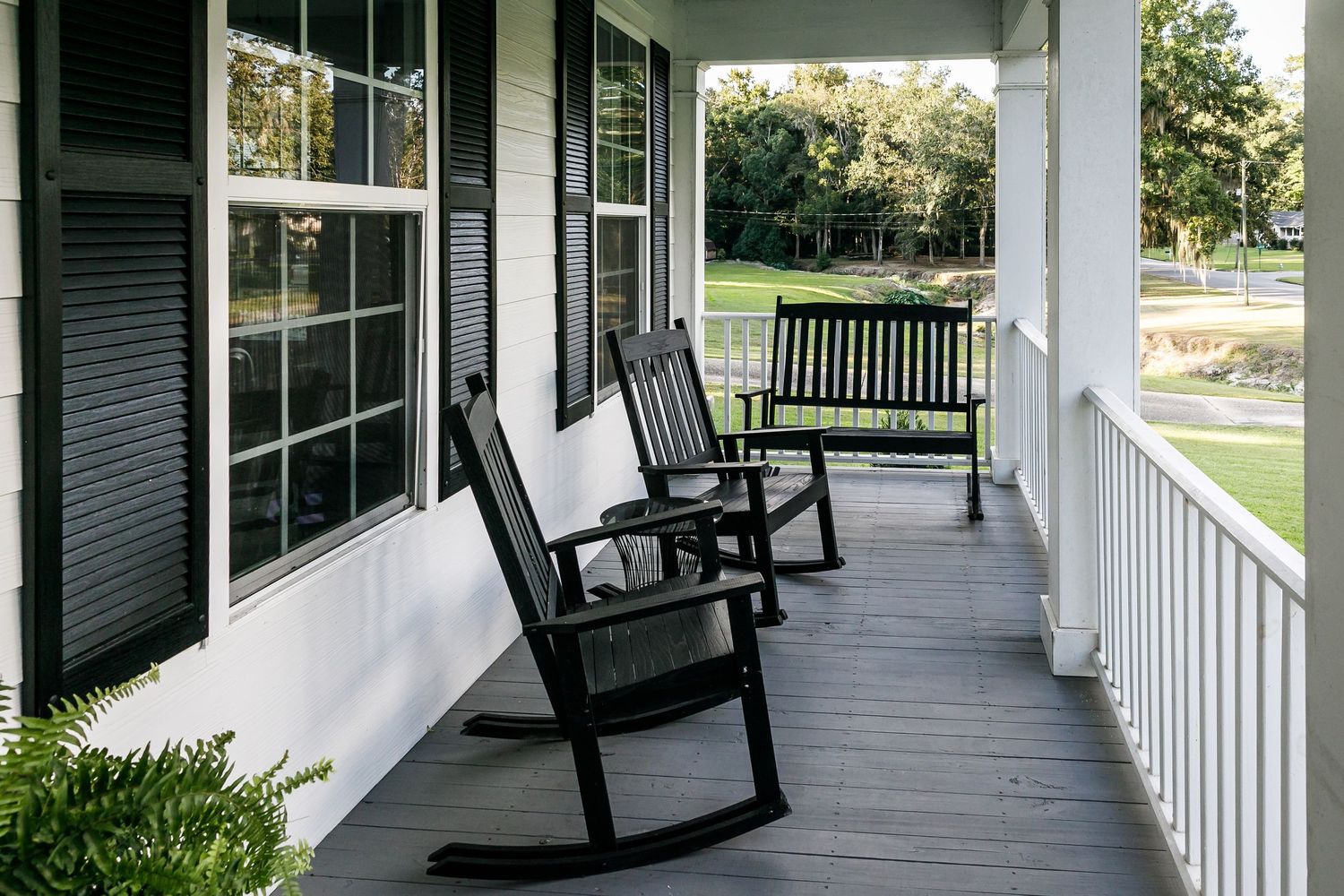 Covered front porch with two black rocking chairs and a bench overlooking a green lawn and trees.