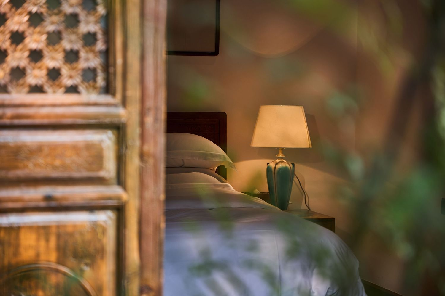 Cozy bedroom corner with a lit bedside lamp, soft pillows and a carved wooden lattice door in warm, dappled light.
