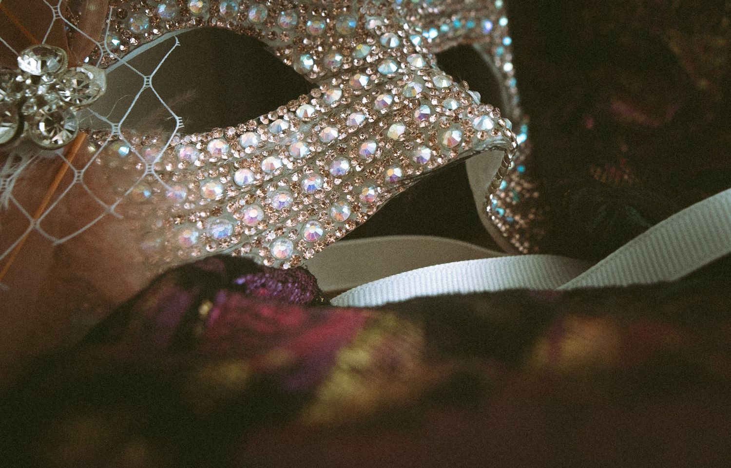Close-up of a bejeweled masquerade mask with sparkling rhinestones and white ribbon on dark fabric.