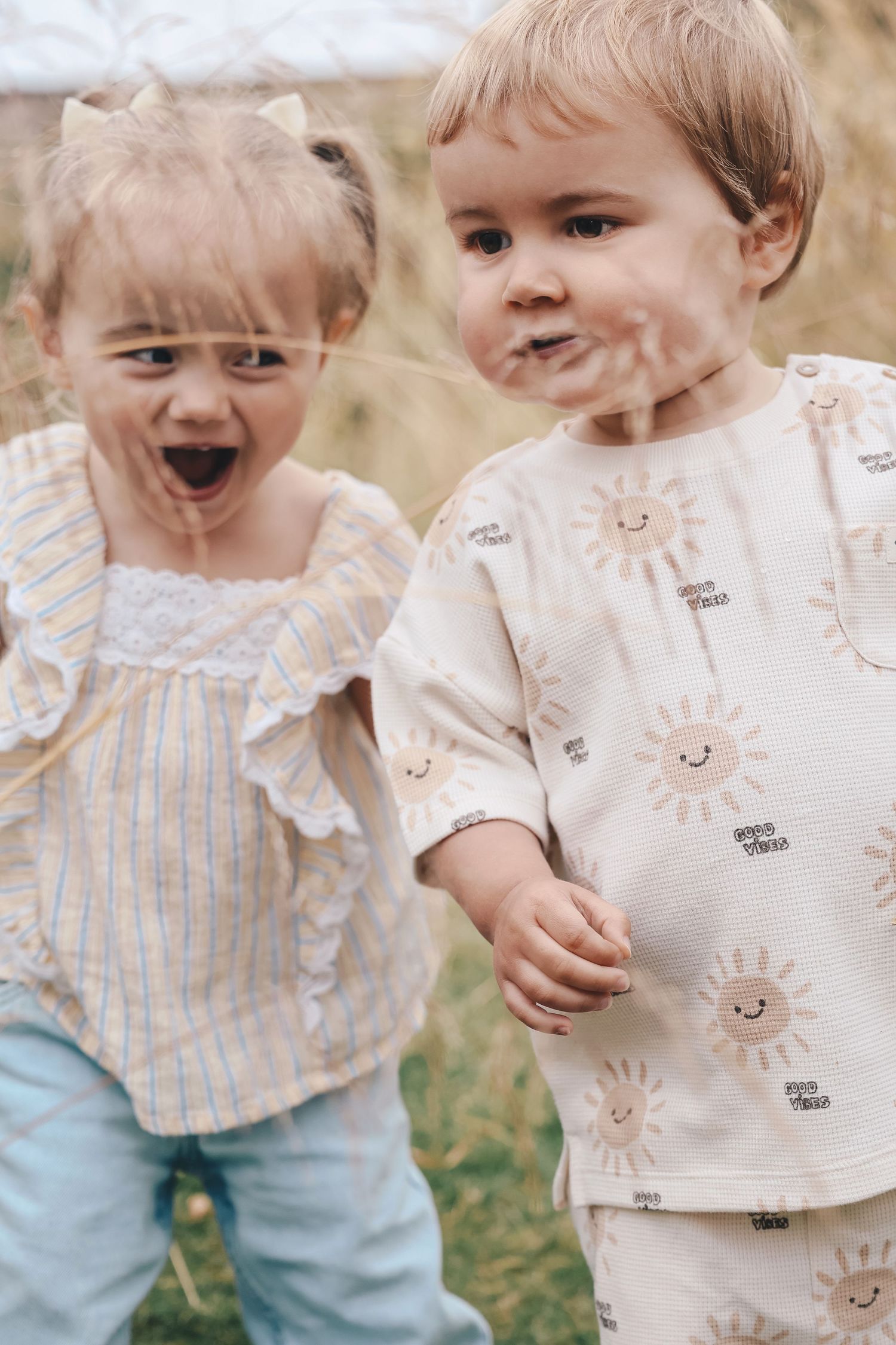 Two toddlers outdoors, girl in striped top with bow, boy in cream sun-print outfit, both with dirt on faces.